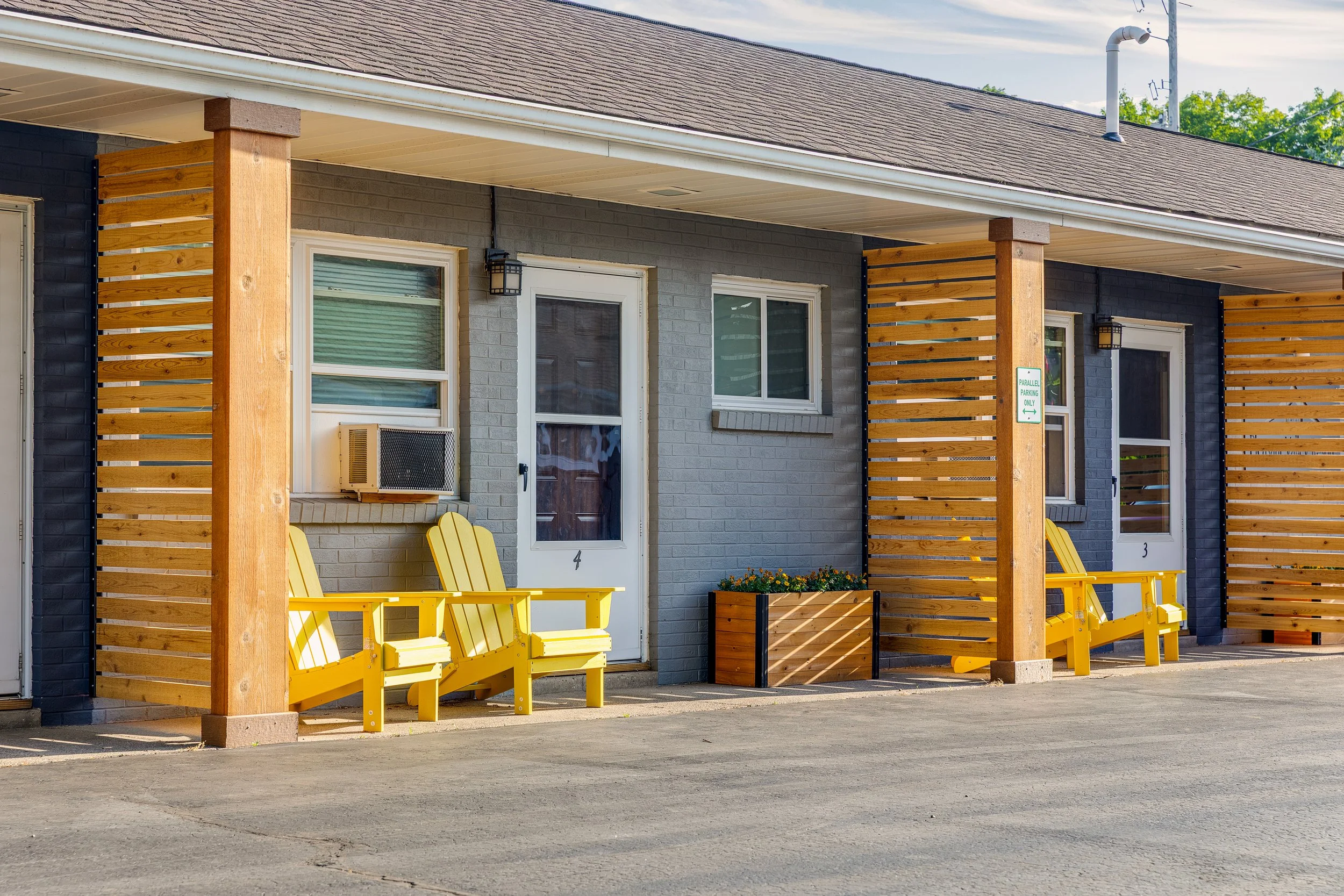 Exterior view of apartments with yellow chairs outside two doors, gray and black brick walls, wooden decorative panels, and small window air conditioner units, on a paved surface.