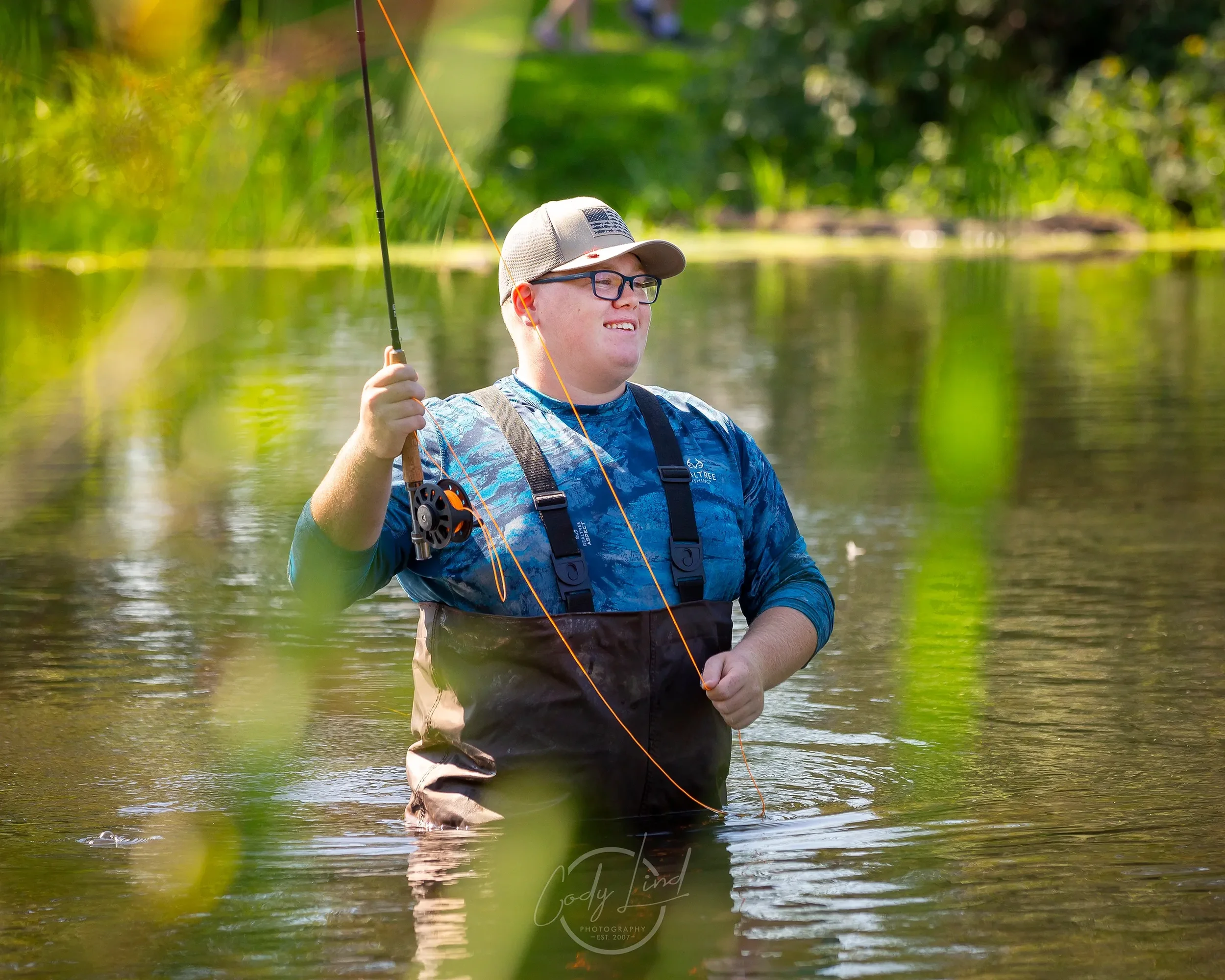 A man standing in a river fishing, wearing a blue shirt, glasses, a gray cap, and waders, holding a fishing rod, with greenery and water reflections in the background.