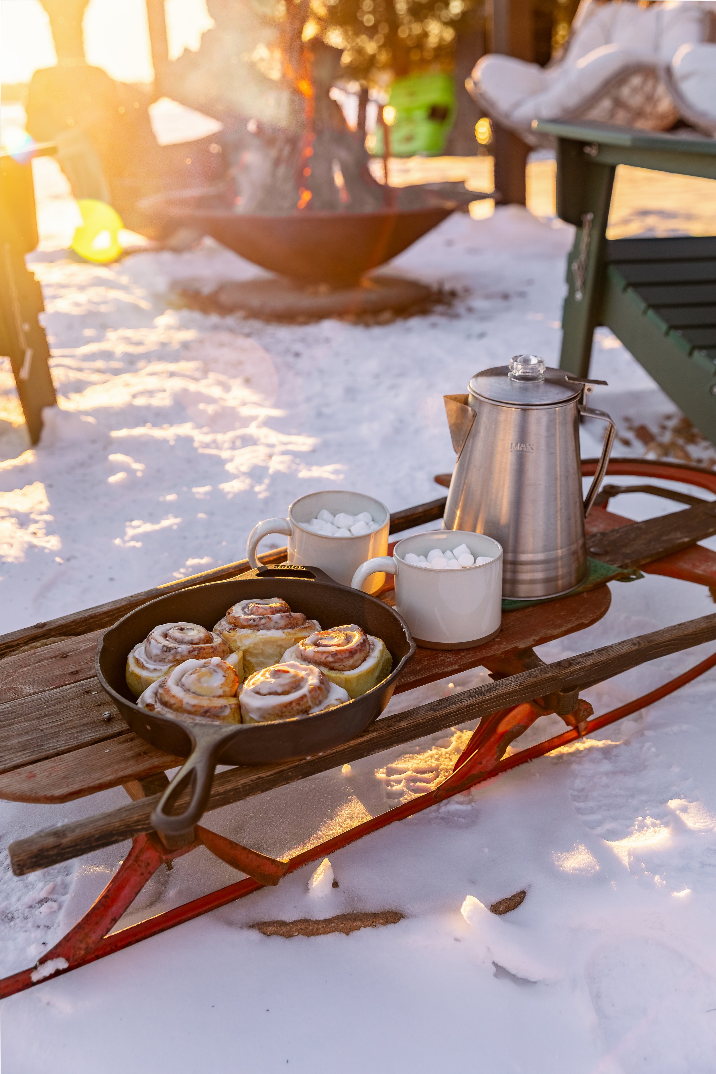 Outdoor scene with a wooden sled carrying cinnamon rolls, a teapot, and two cups filled with marshmallows on a snowy surface during sunset.
