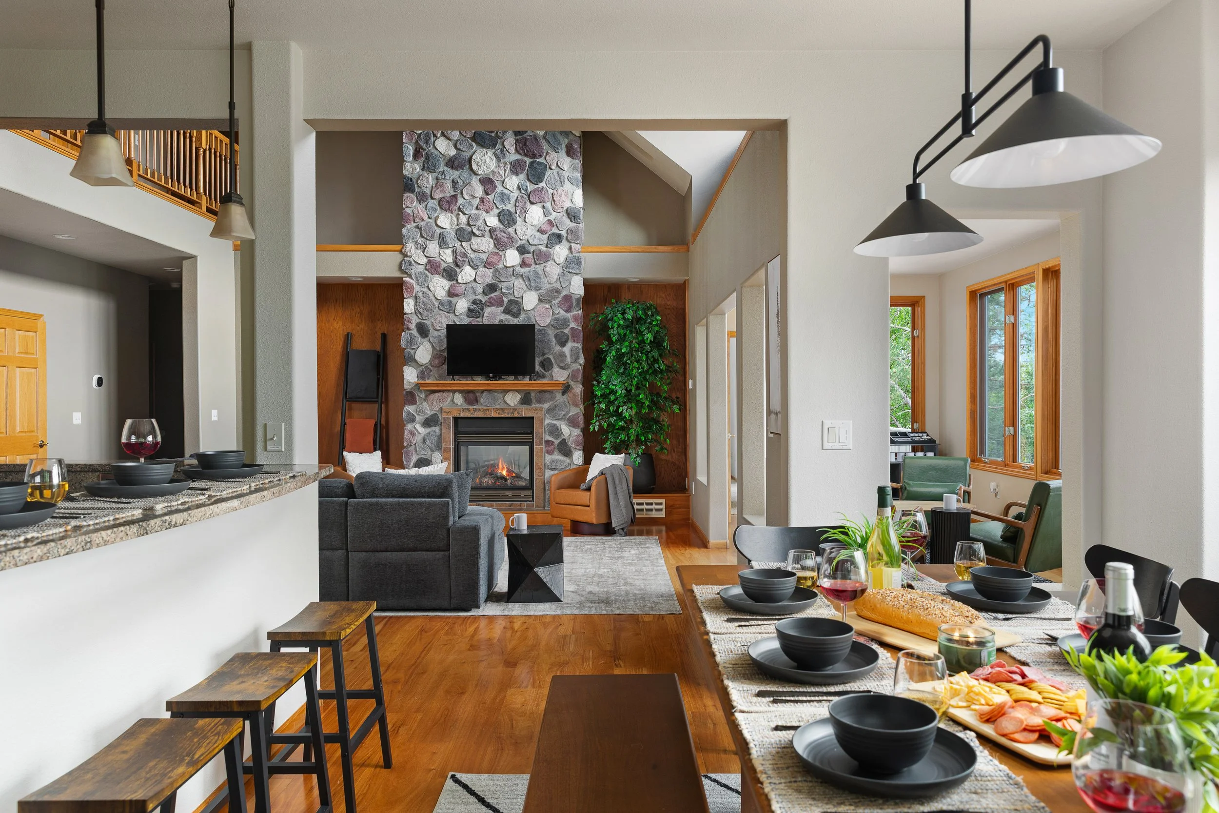 Open-concept living and dining room with wooden floors, a stone fireplace, a mounted TV, a gray sofa, and a dining table set for a meal with black dishware, wine glasses, and food, in a house with large windows.