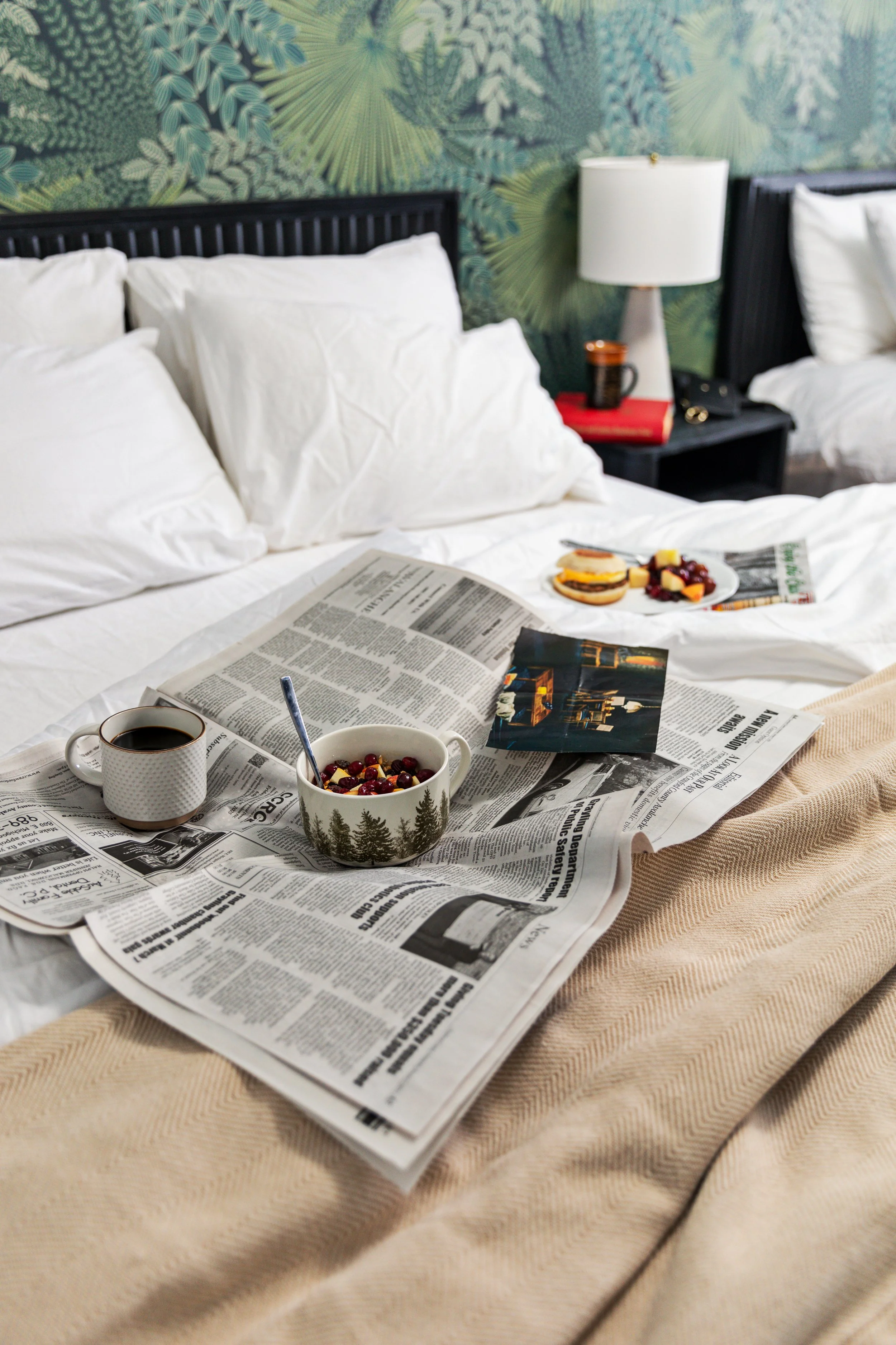 A cozy bed with white sheets and pillows, a newspaper spread out on the bed, a cup of coffee, a bowl of berries with a spoon, a small photo, and breakfast foods including a muffin and fruit on the bed. There are lamps and bedside tables in the backgr
