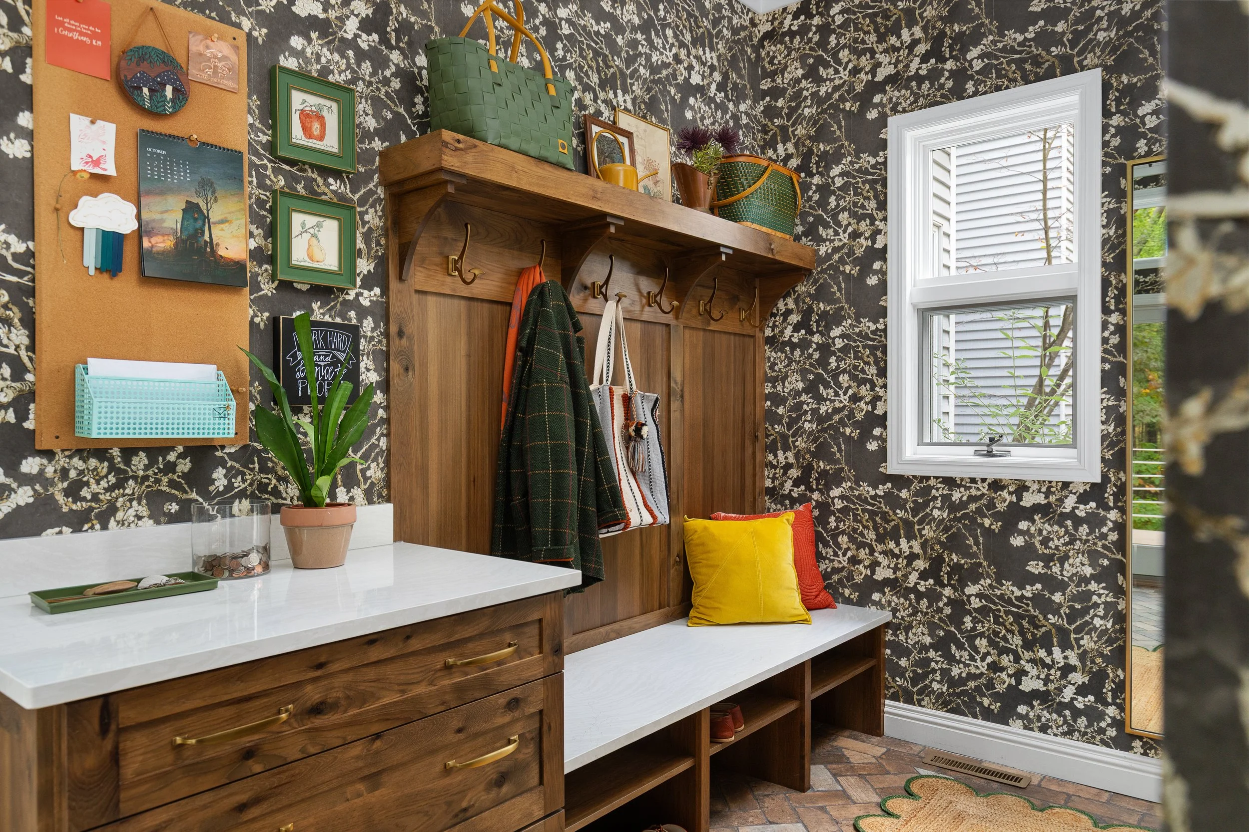 Entryway with a wooden bench, yellow and red pillows, coat hooks with hanging coats and bags, a shelf with baskets and decorative items, a potted plant on a white countertop, a bulletin board with notes and artwork, a window with white trim showing a