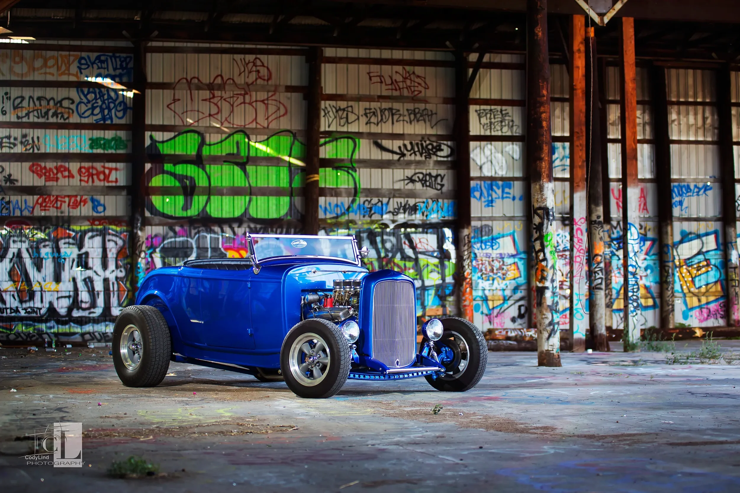 A classic blue hot rod car with no roof in an abandoned building with graffiti-covered walls.
