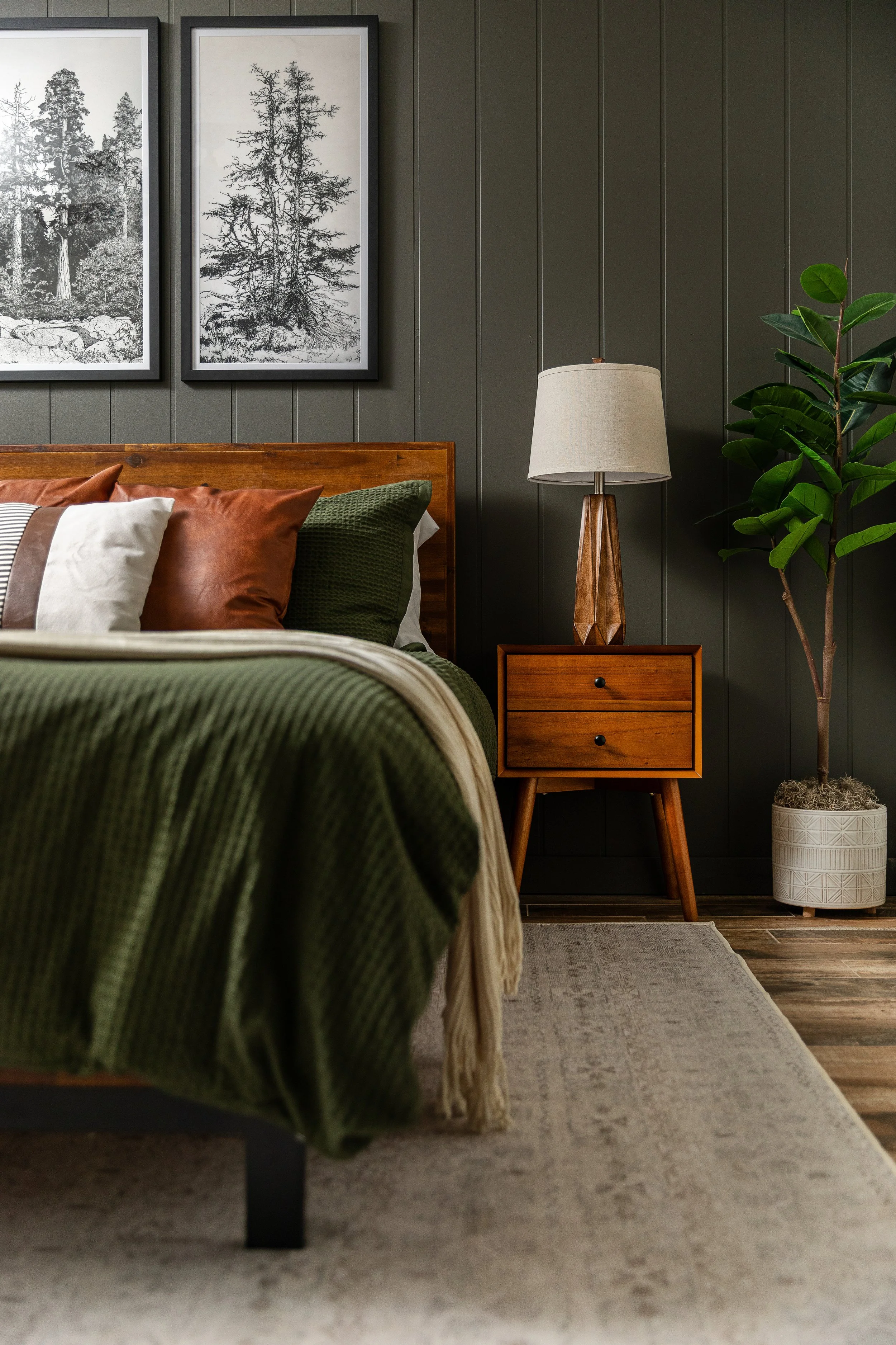 A bedroom with a green bedspread, decorative pillows, a wooden headboard, a nightstand with a lamp, framed black-and-white forest photos, a potted plant, and a grey rug on a wood floor.