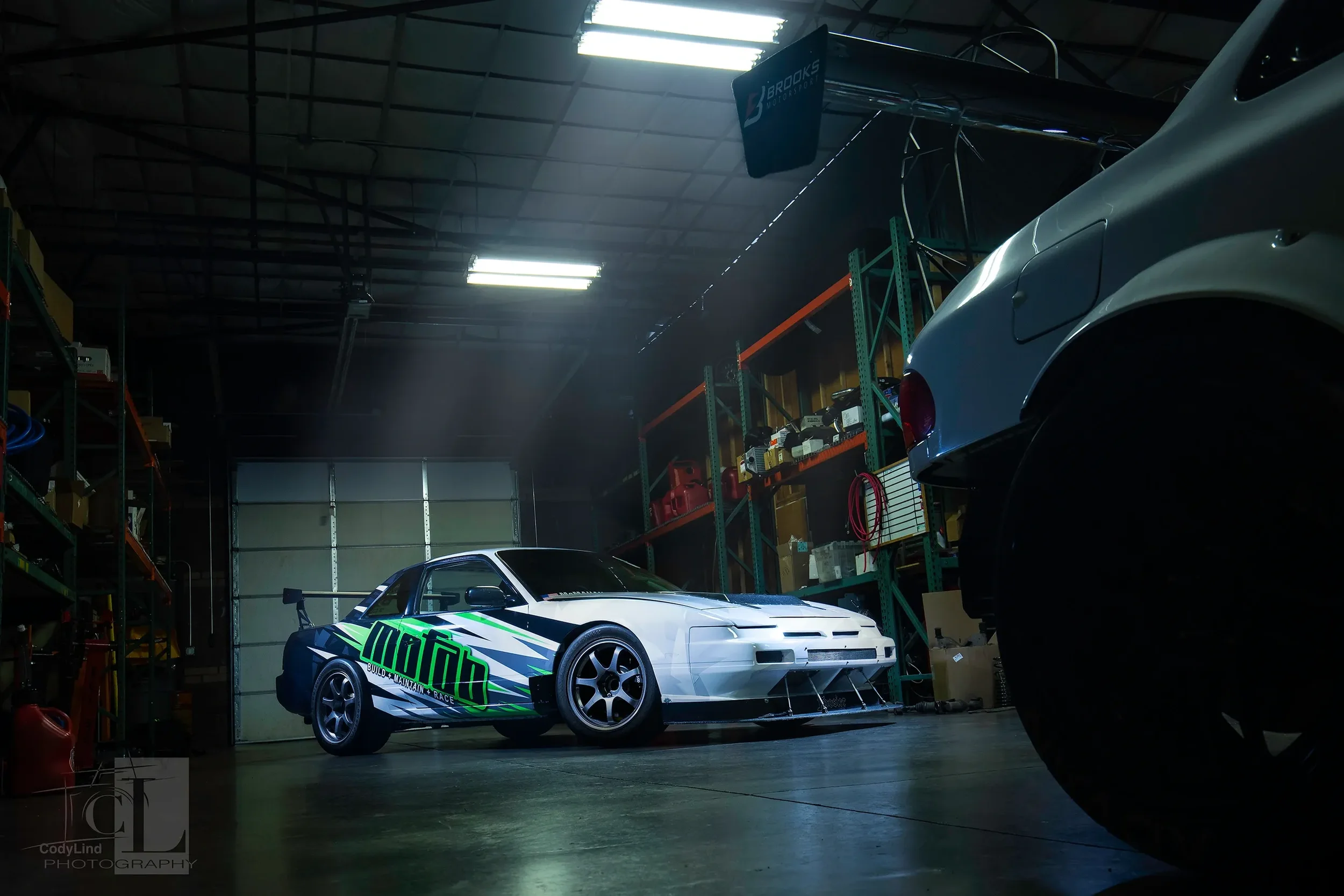 Inside a garage with race cars, tools, and equipment on shelves. A race car with a green and black design is in the center, with another vehicle partially visible on the right.
