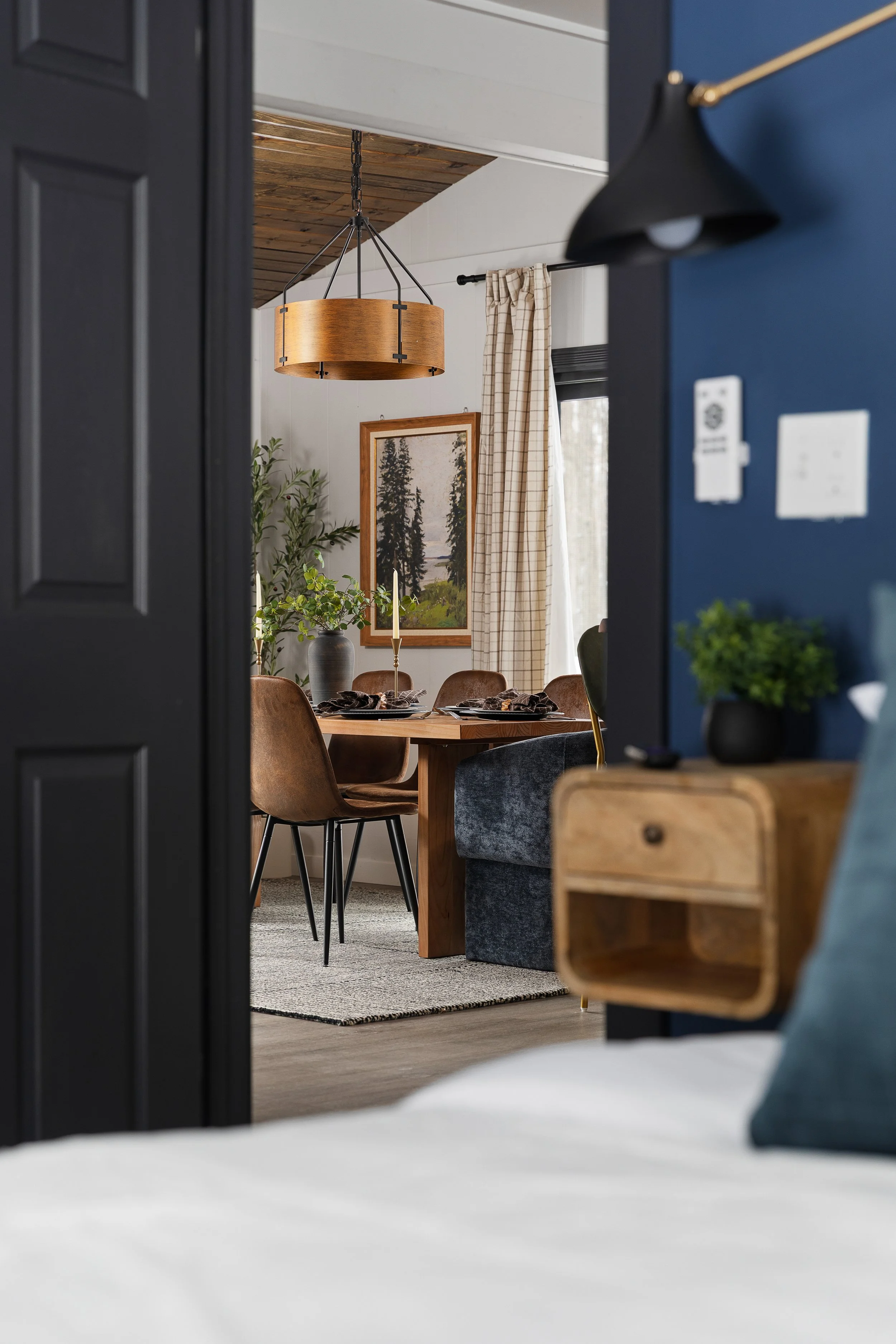 View through a doorway into a dining room with a wooden table, six chairs, a large landscape painting on the wall, a tall plant, and a window with checked curtains.