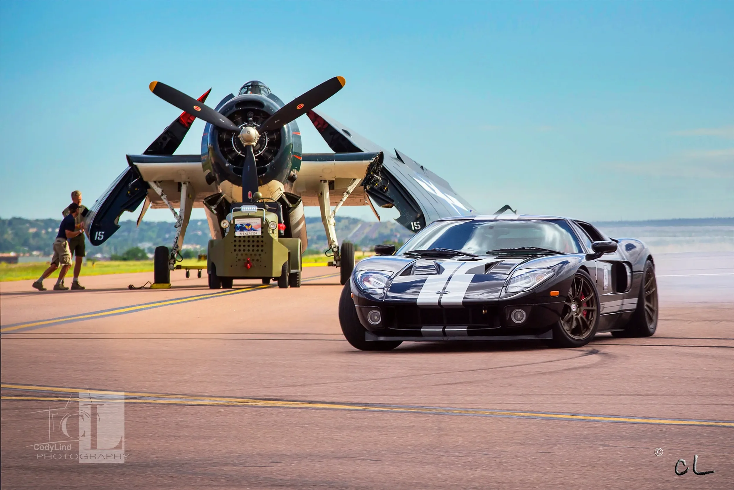 A black sports car with white racing stripes parked on an airstrip with a vintage military aircraft in the background, and two people walking nearby.