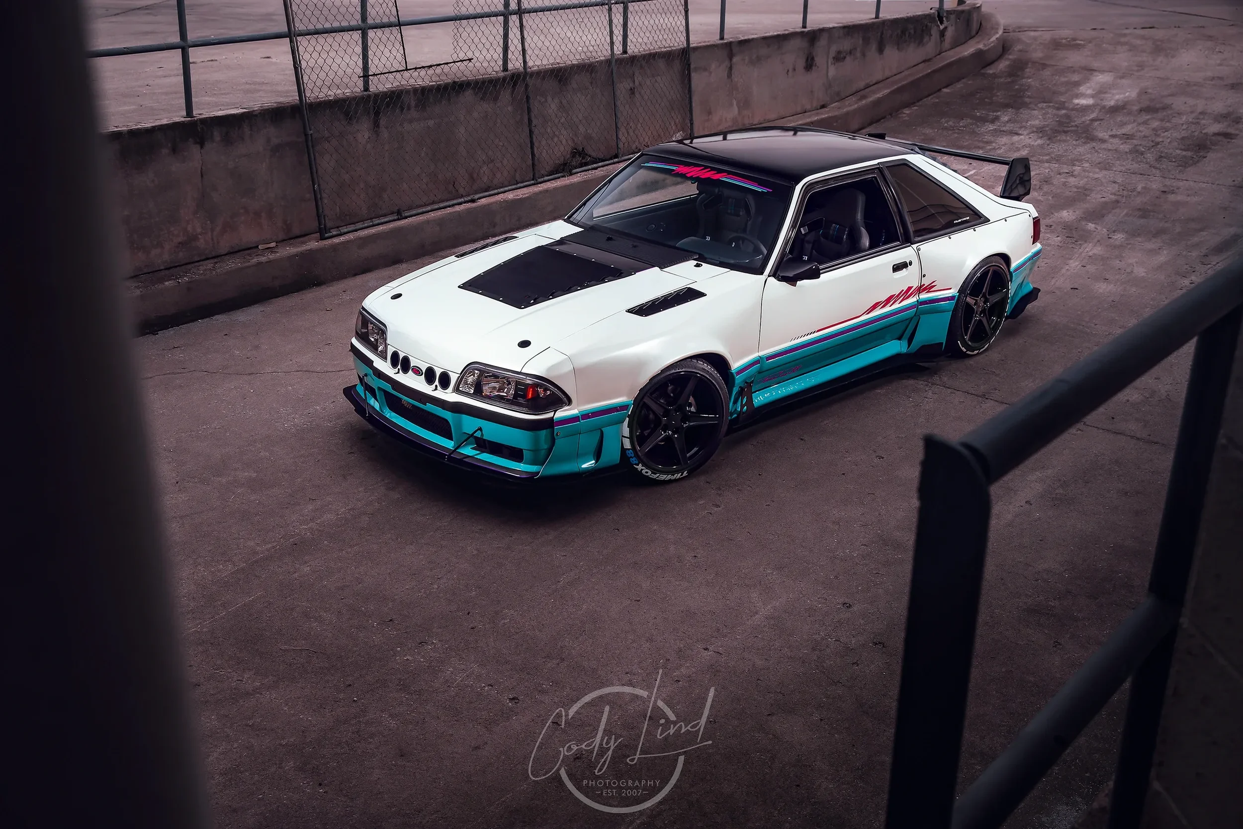 A custom white and turquoise race car with black accents and graphics, parked on a concrete surface near a chain-link fence.