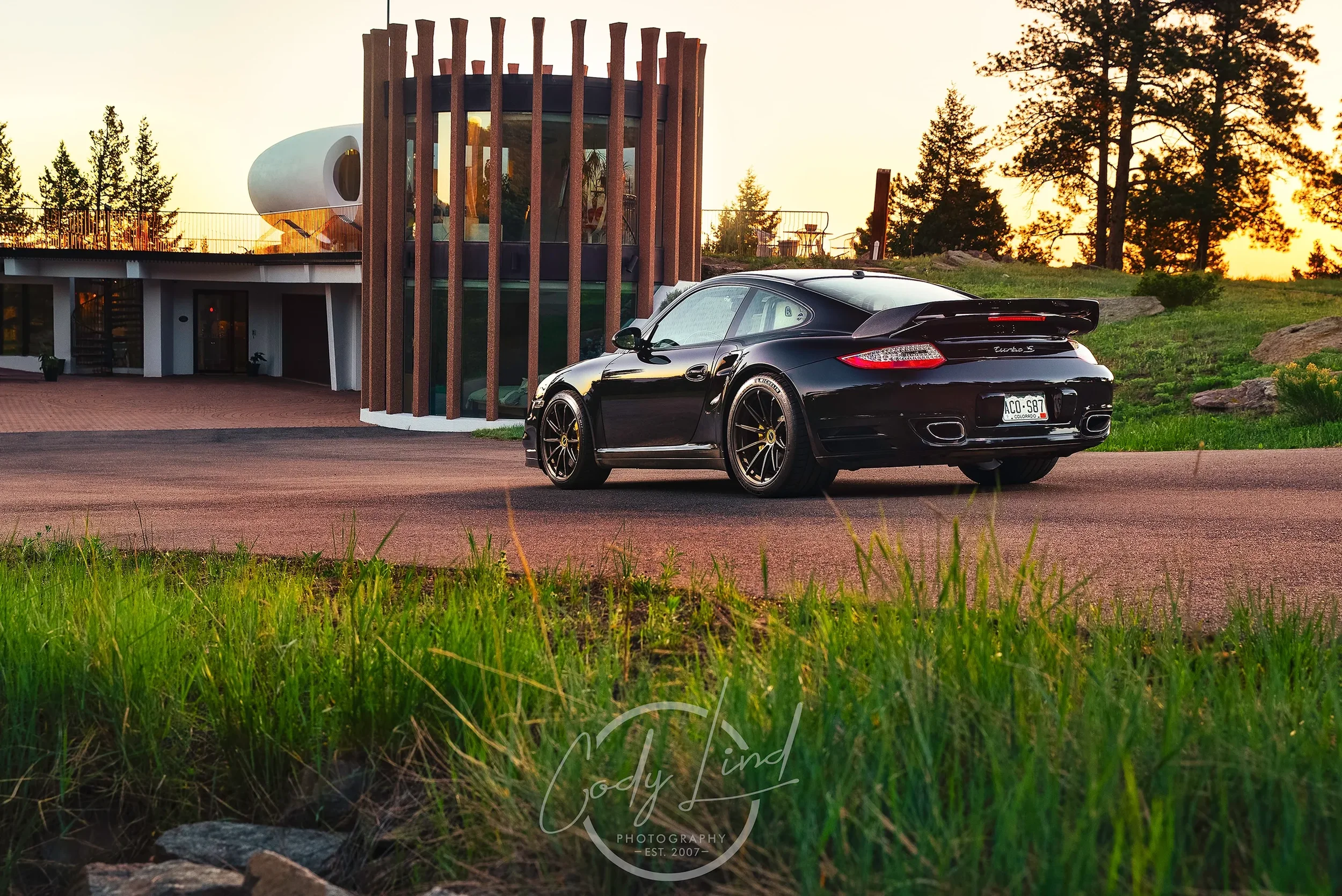 A black Porsche 911 parked in front of a modern building with rounded design elements, wooden vertical slats, and large glass windows, at sunset with trees in the background and green grass in the foreground.