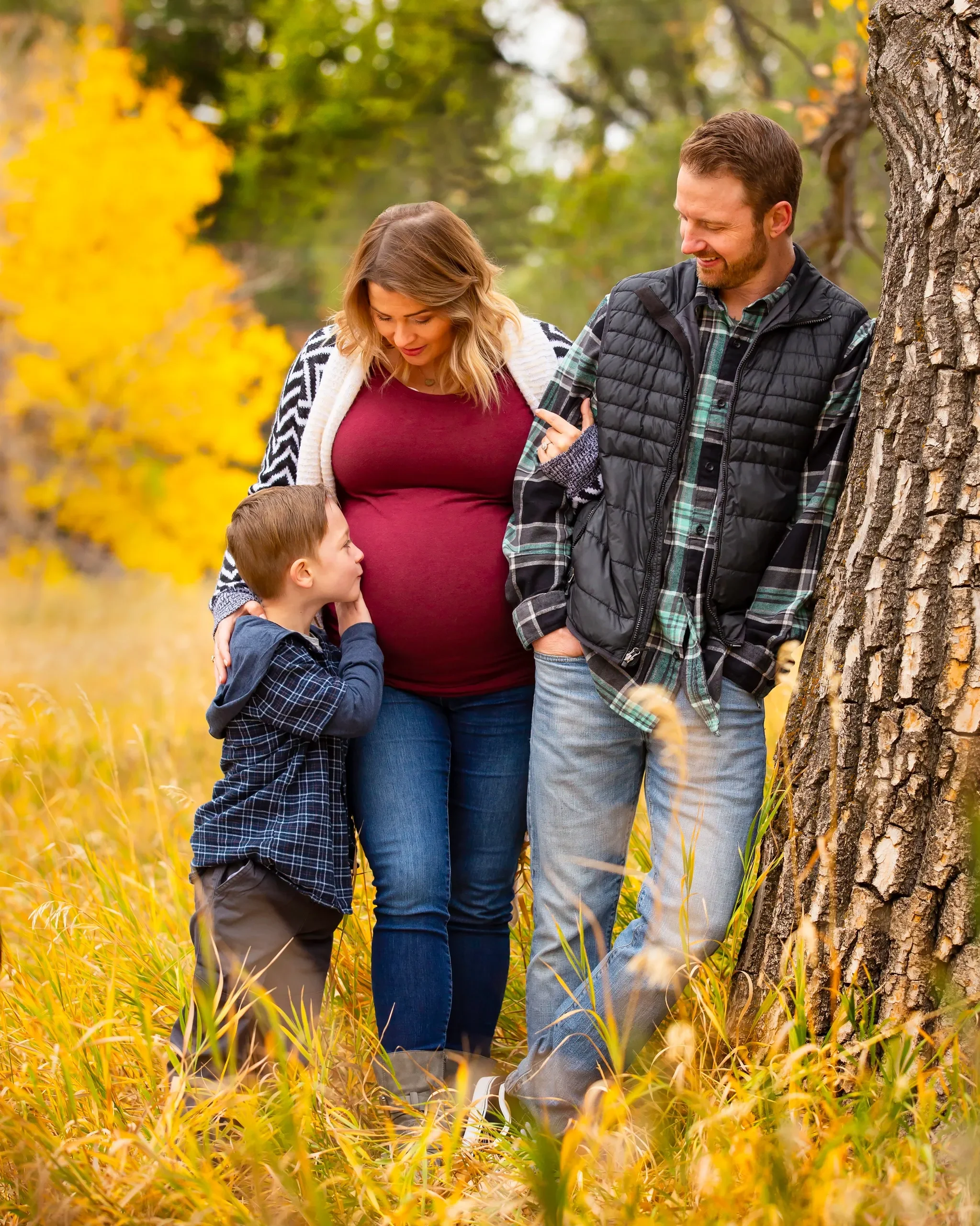 Family of three enjoying autumn outside; mother, father, and young son standing next to a tree surrounded by yellow fall leaves.