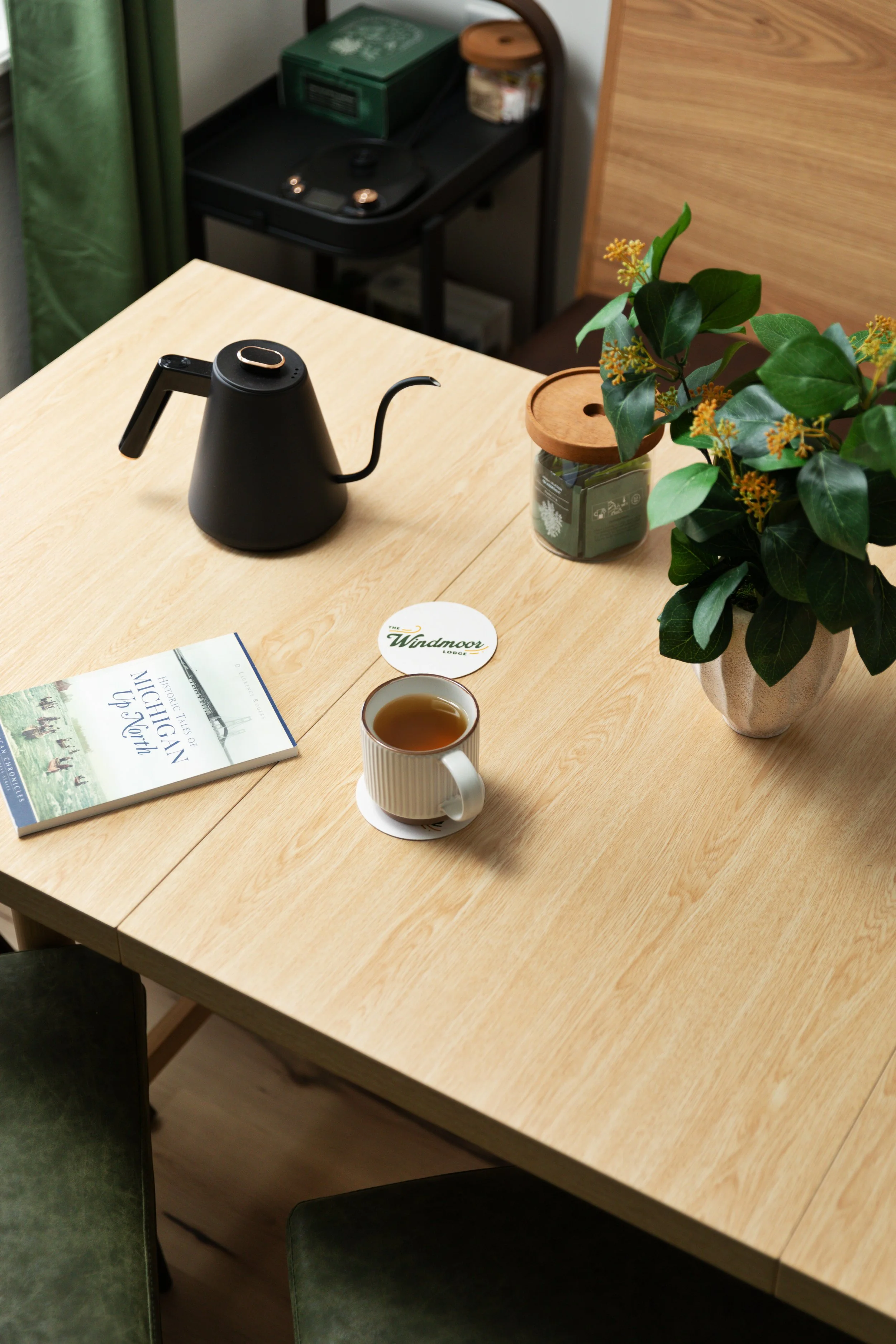 A wooden table with a cup of tea, a magazine about Michigan, a plant in a silver pot, a black electric kettle, and a coaster with the word "Windmoor" on it.