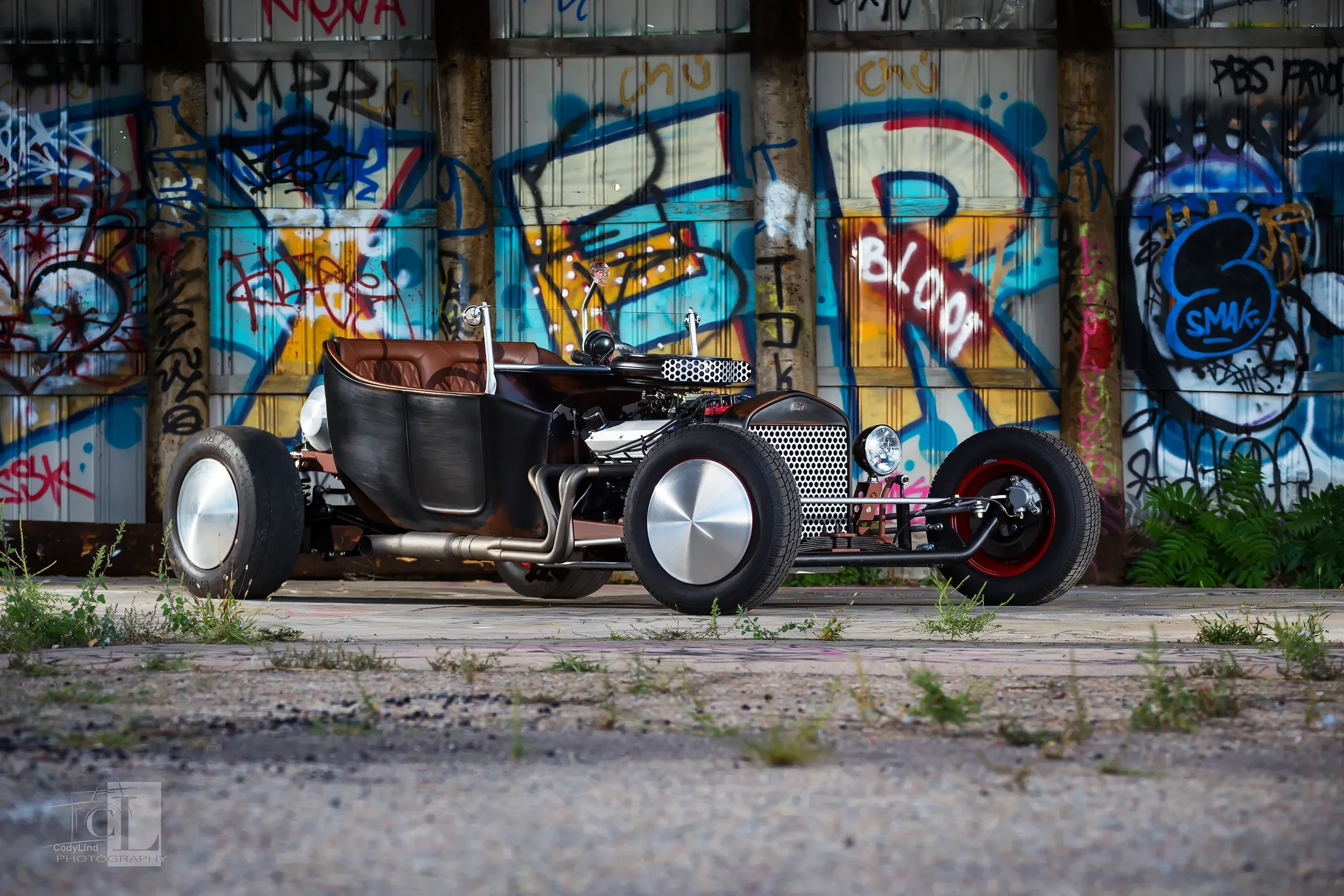 A vintage-style hot rod car with a black body, brown leather seats, and large silver hubcap wheels, parked in front of a graffiti-covered metal wall.