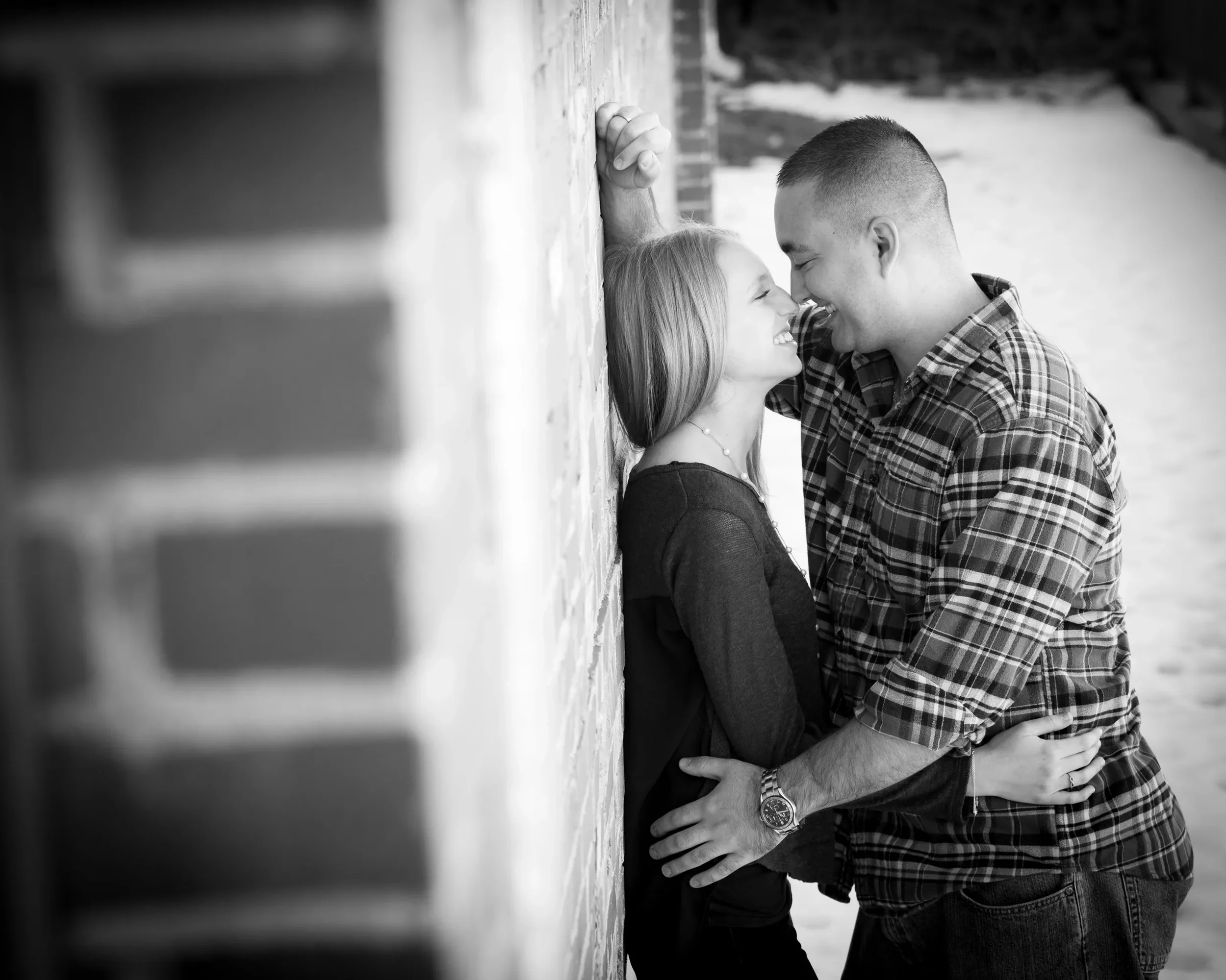 A black and white photo of a couple smiling and touching noses, standing close against a brick wall. The woman has long hair and is wearing a dark top, while the man has short hair, a checked shirt, and is wearing a watch. They seem happy and intimat