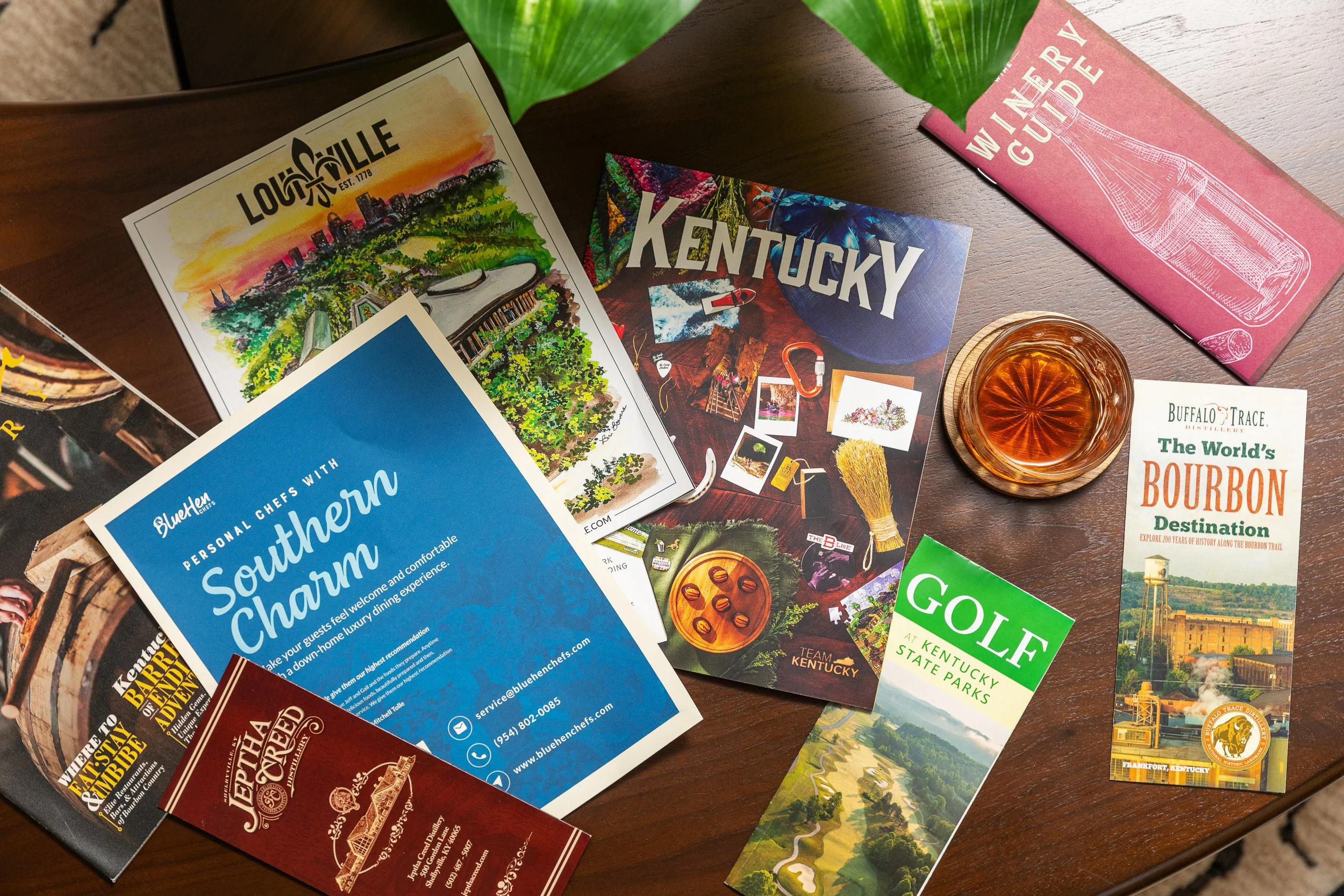 A top view of a table with various Kentucky travel brochures, a glass of bourbon, and related promotional materials.