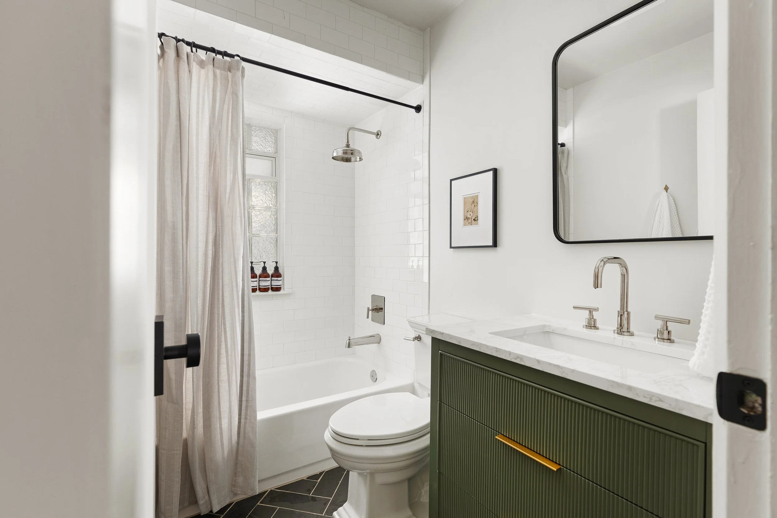 A modern bathroom with a white bathtub and shower, a green vanity with a white marble countertop, a rectangular mirror, and a small framed picture on the wall.