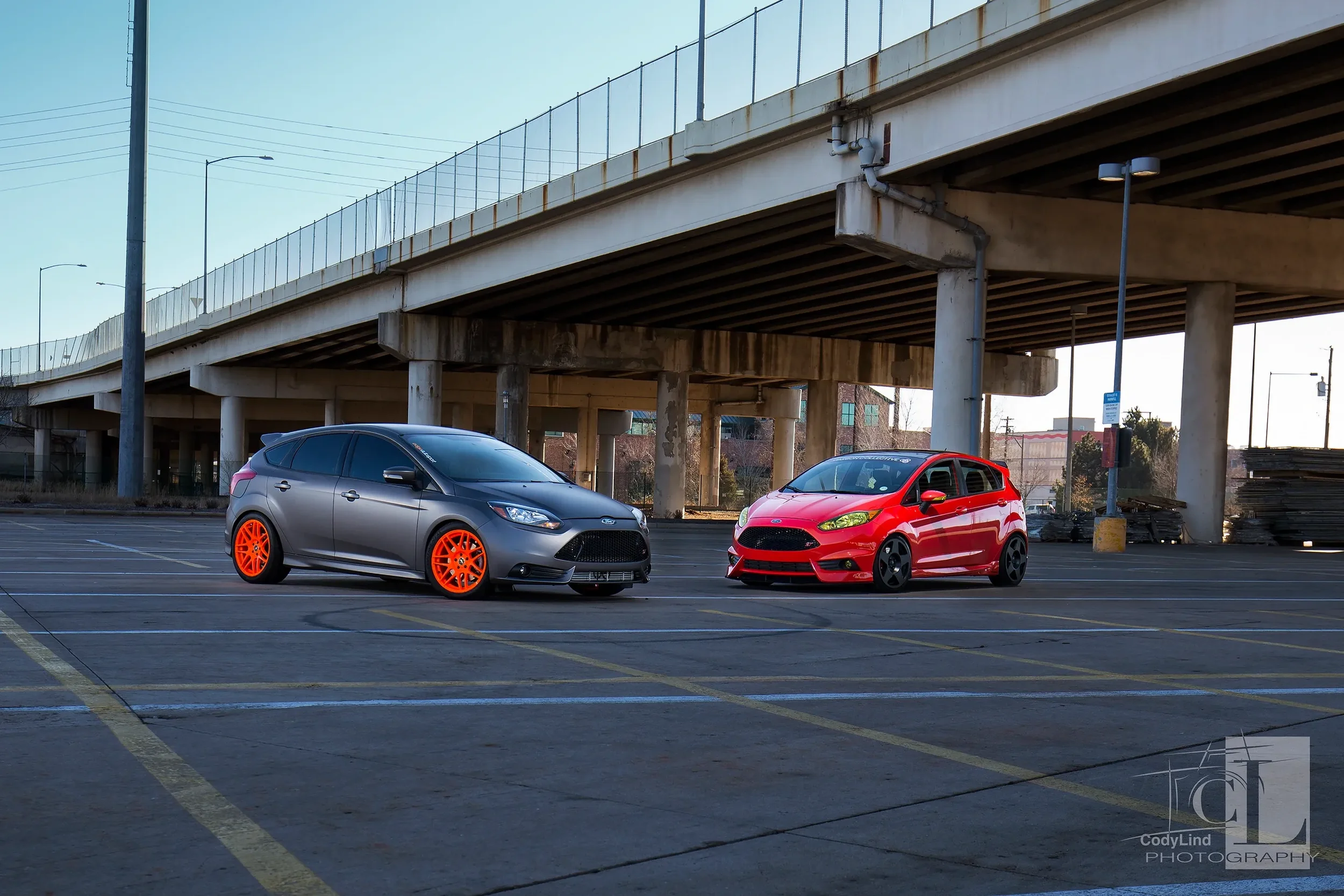 Two parked cars, a gray car with bright orange wheels and a red car with black wheels, under an overpass in a mostly empty parking lot.