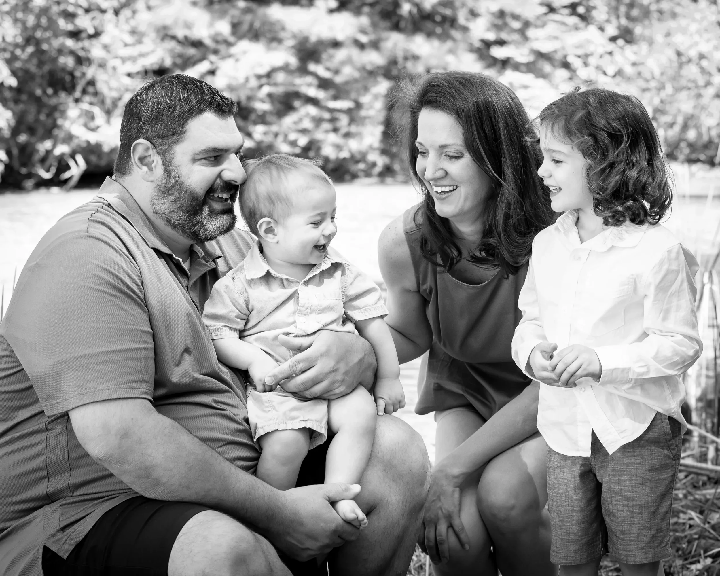 A family of four, including two adults and two children, enjoying a happy moment outdoors near a body of water with trees in the background. They are smiling and looking at each other.