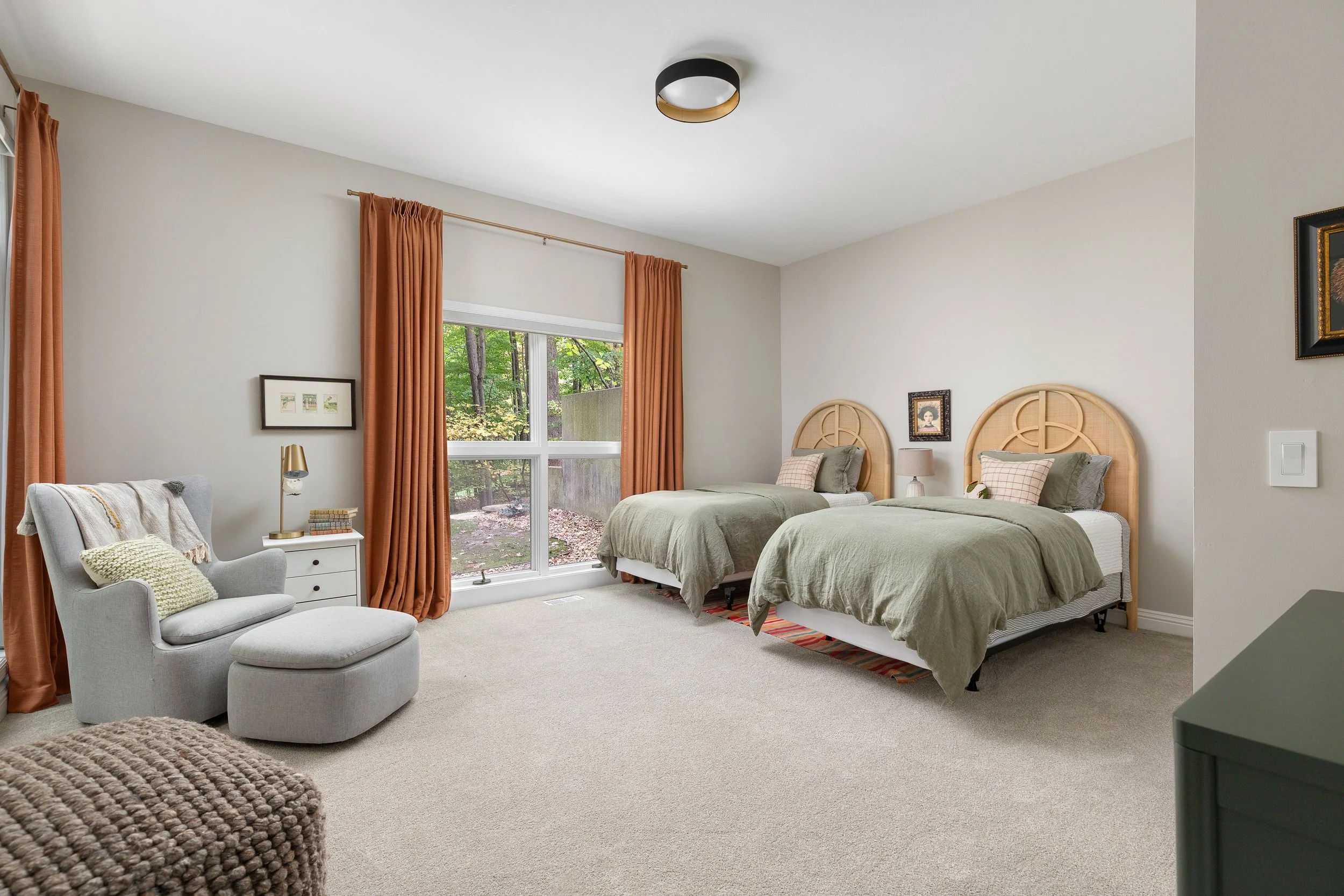 A bedroom with two twin beds with rattan headboards, green bedding, and pillows, a window with orange curtains, a gray chair with a matching ottoman, a white side table, and decorative artwork on the walls.