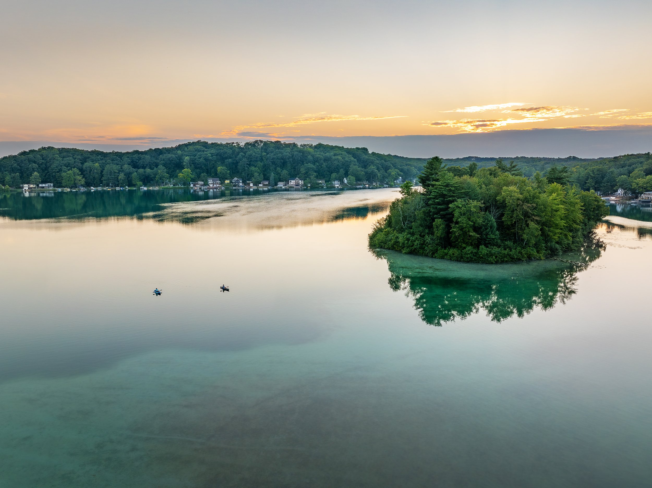 A peaceful lake scene at sunset with an island covered in trees near the right side, two kayakers paddling on the calm water, and a tree-lined shoreline with houses in the background.