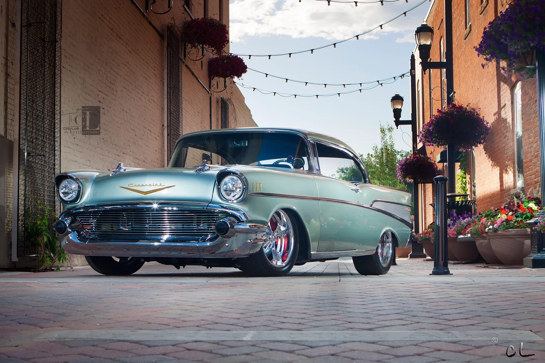 A vintage Chevrolet car parked on a brick street alley with hanging string lights, flower pots, and brick buildings on both sides.