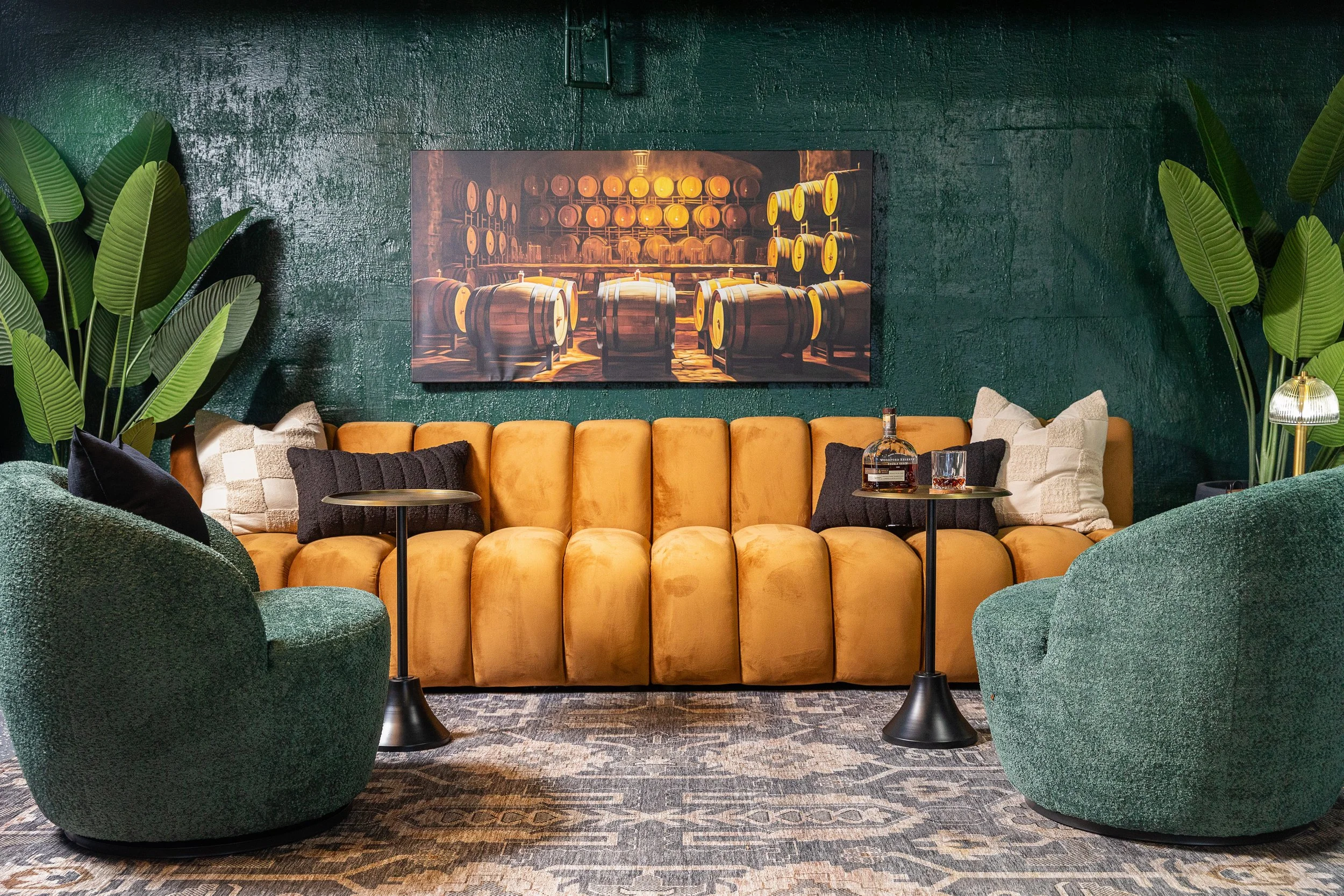 Living room with a mustard yellow sofa, two green armchairs, potted plants, wall art of wine barrels, and side tables with drinks.