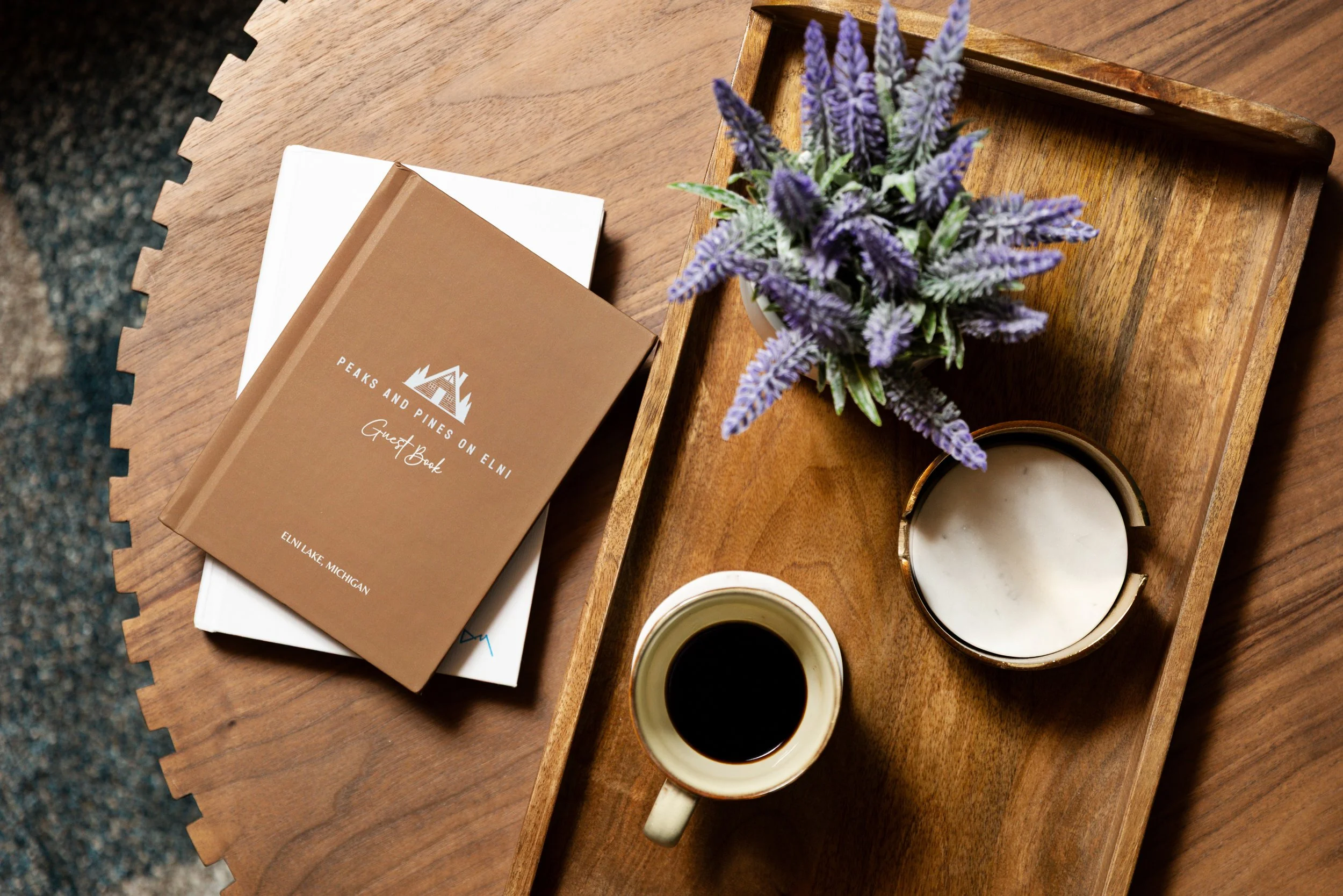 A wooden tray on a table holding a potted purple flowering plant, a cup of black coffee, and a white candle, with a guest book and a white notebook nearby.
