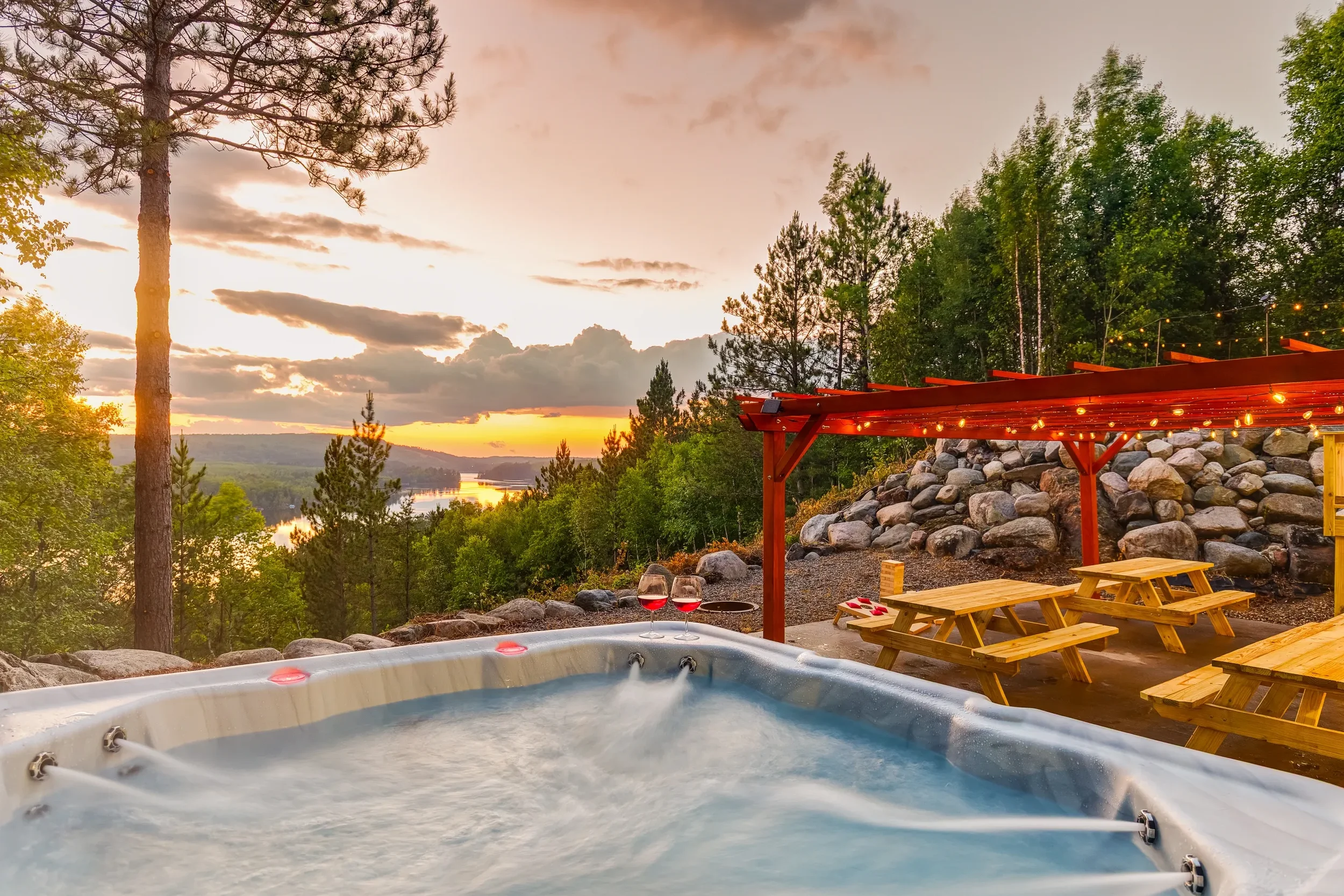 An outdoor hot tub with two glasses of red wine on its edge, surrounded by a wooden patio with picnic tables, a wooden pergola with string lights, and a scenic view of a river and hills at sunset, with trees and clouds in the sky.