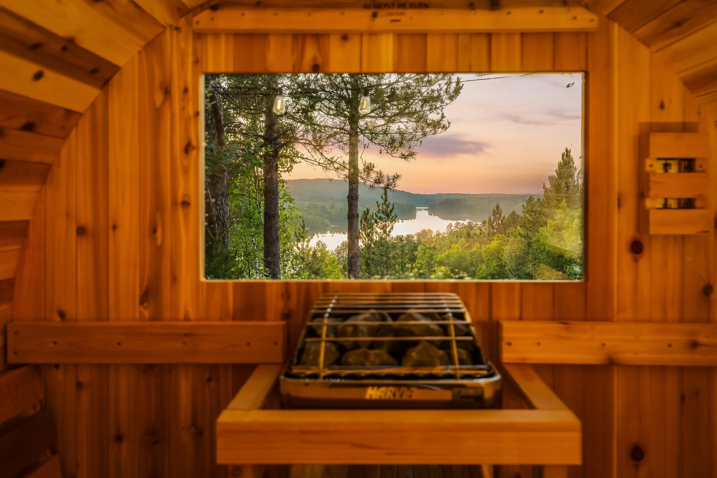 View of a river and trees through a large window in a wooden sauna, with a sauna heater filled with rocks in the foreground.