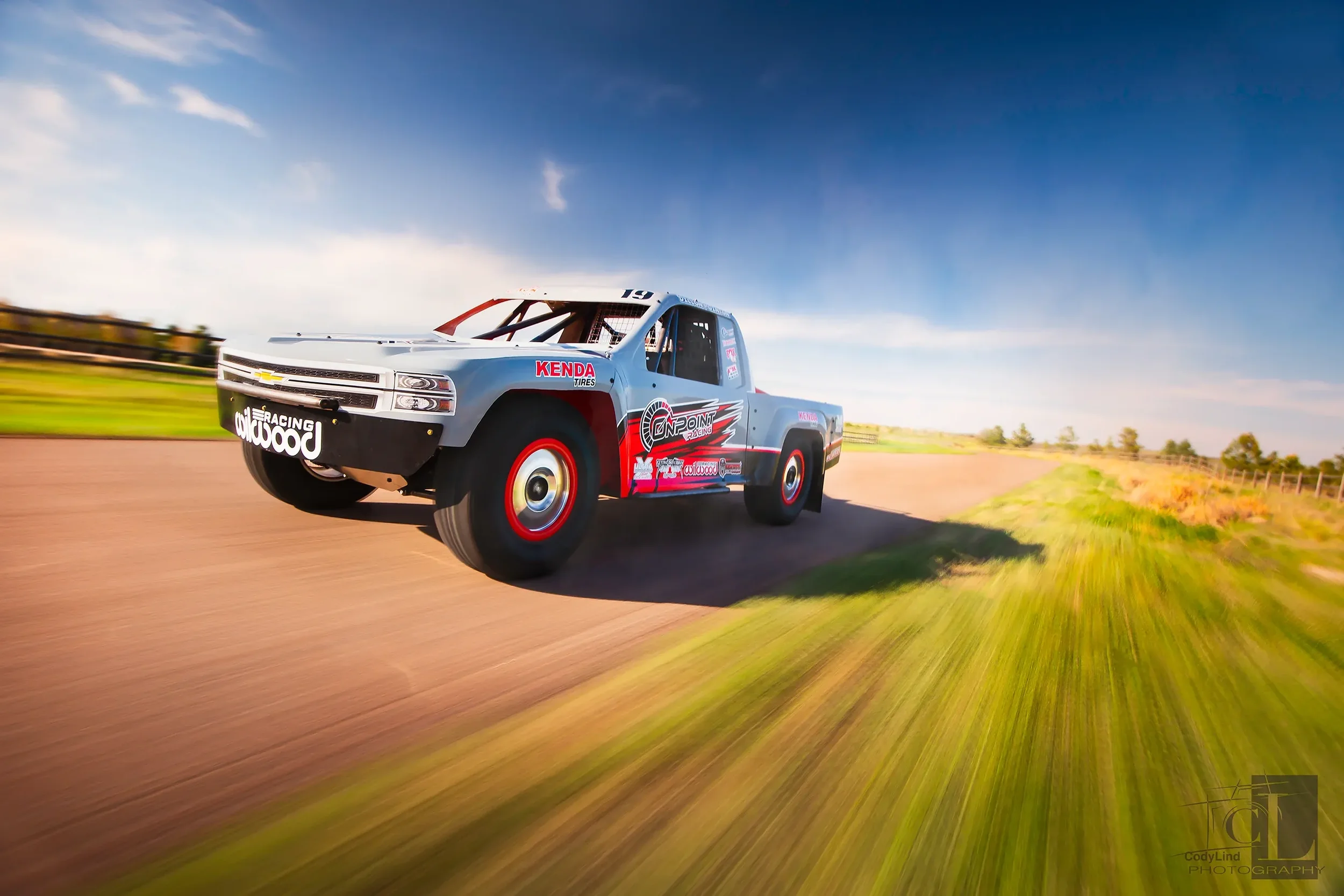 Racing truck driving on a dirt track with motion blur and a blue sky in the background.