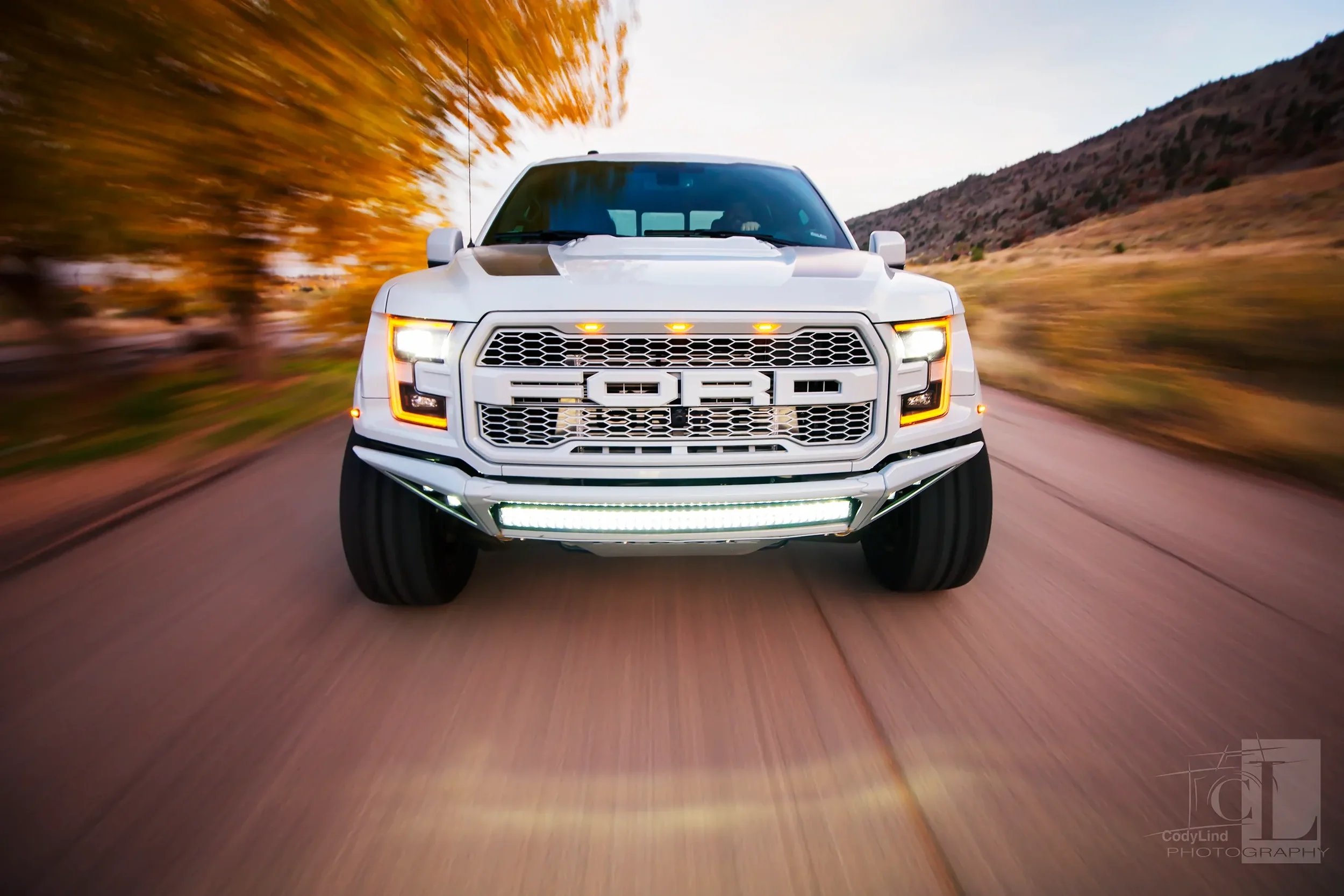 White Ford pickup truck driving on a rural road with autumn trees and hills in the background, shot from a front perspective during dusk.