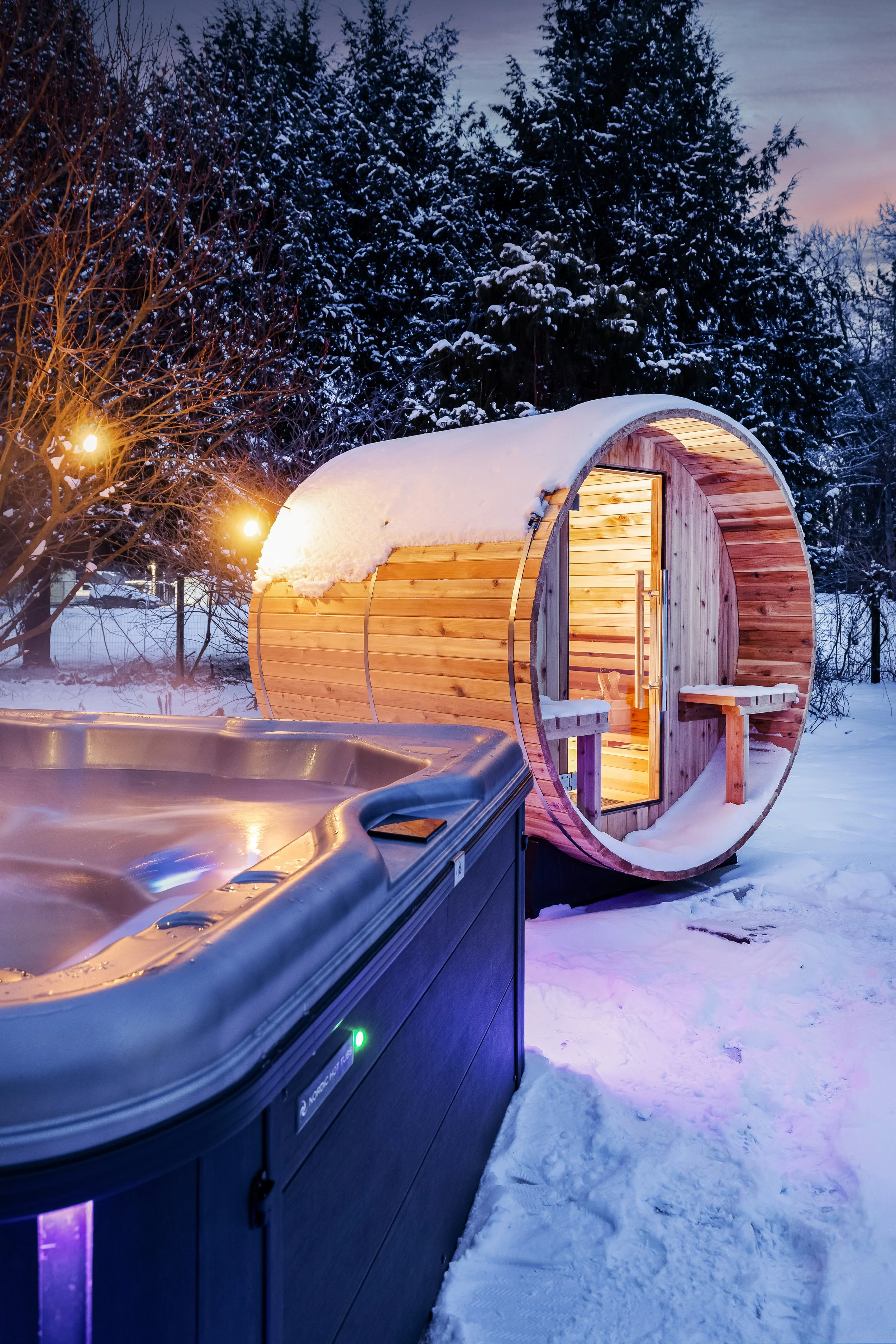 A snow-covered backyard with a wooden barrel-shaped sauna emitting warm light, next to a hot tub, surrounded by snow and trees, at dusk.