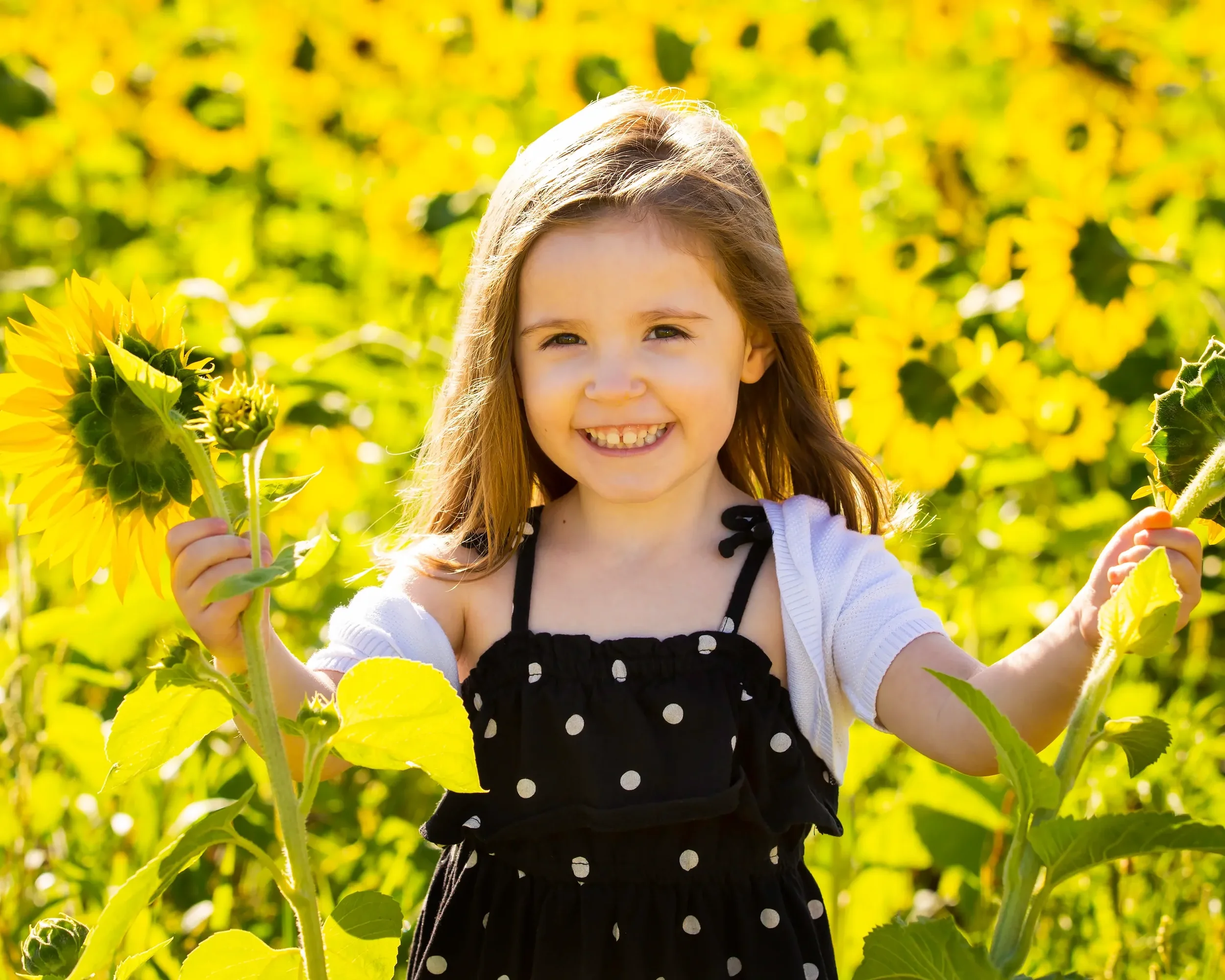 A young girl with long brown hair smiling in a sunflower field, holding sunflower blooms in both hands.