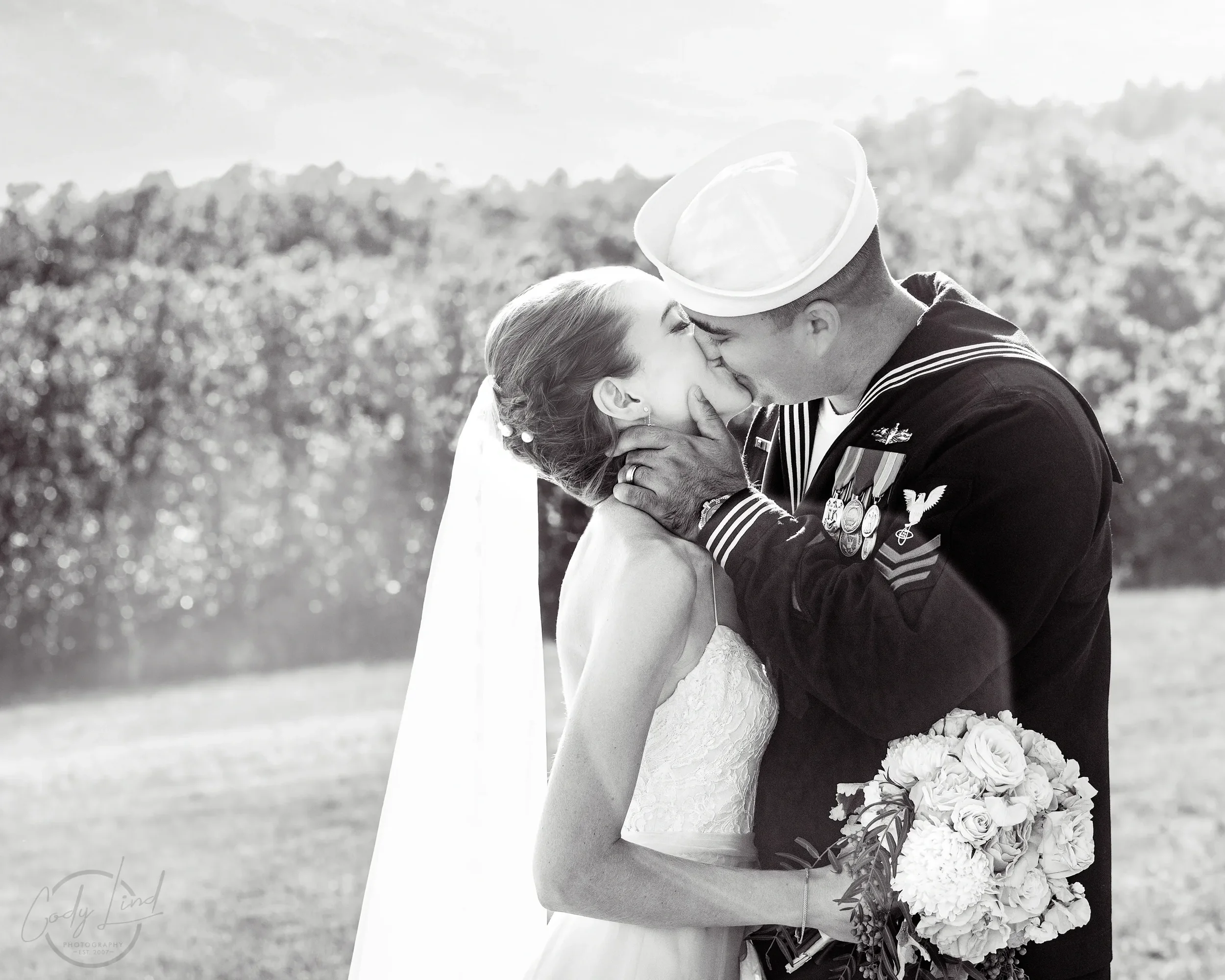 A bride and a groom in military uniform sharing a kiss outdoors, with the bride holding a bouquet of flowers.