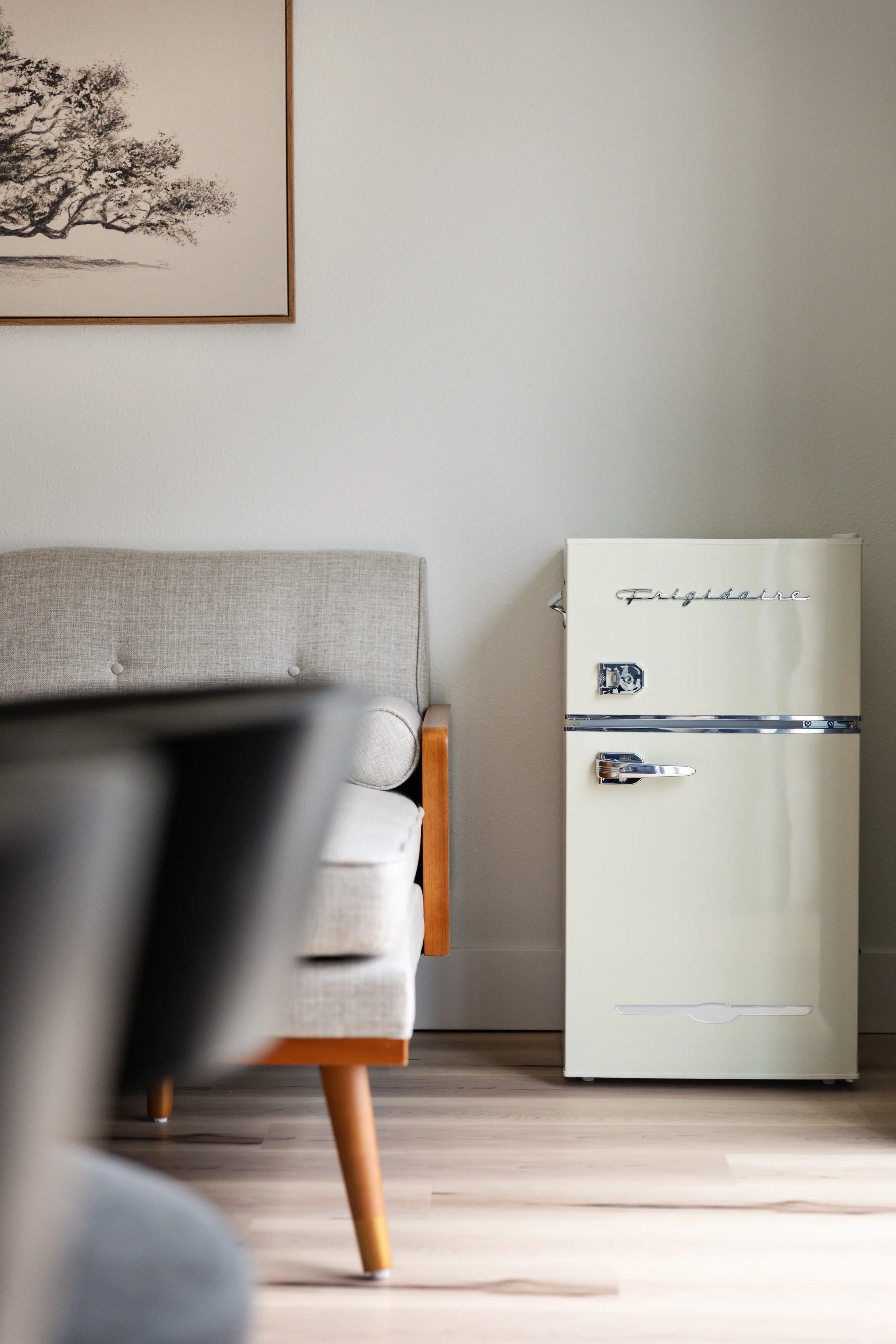 A vintage white Frigidaire refrigerator next to a beige mid-century modern sofa with wooden armrests and tapered wooden legs in a minimalist room with a light-colored wooden floor and a line drawing of a tree on the wall.