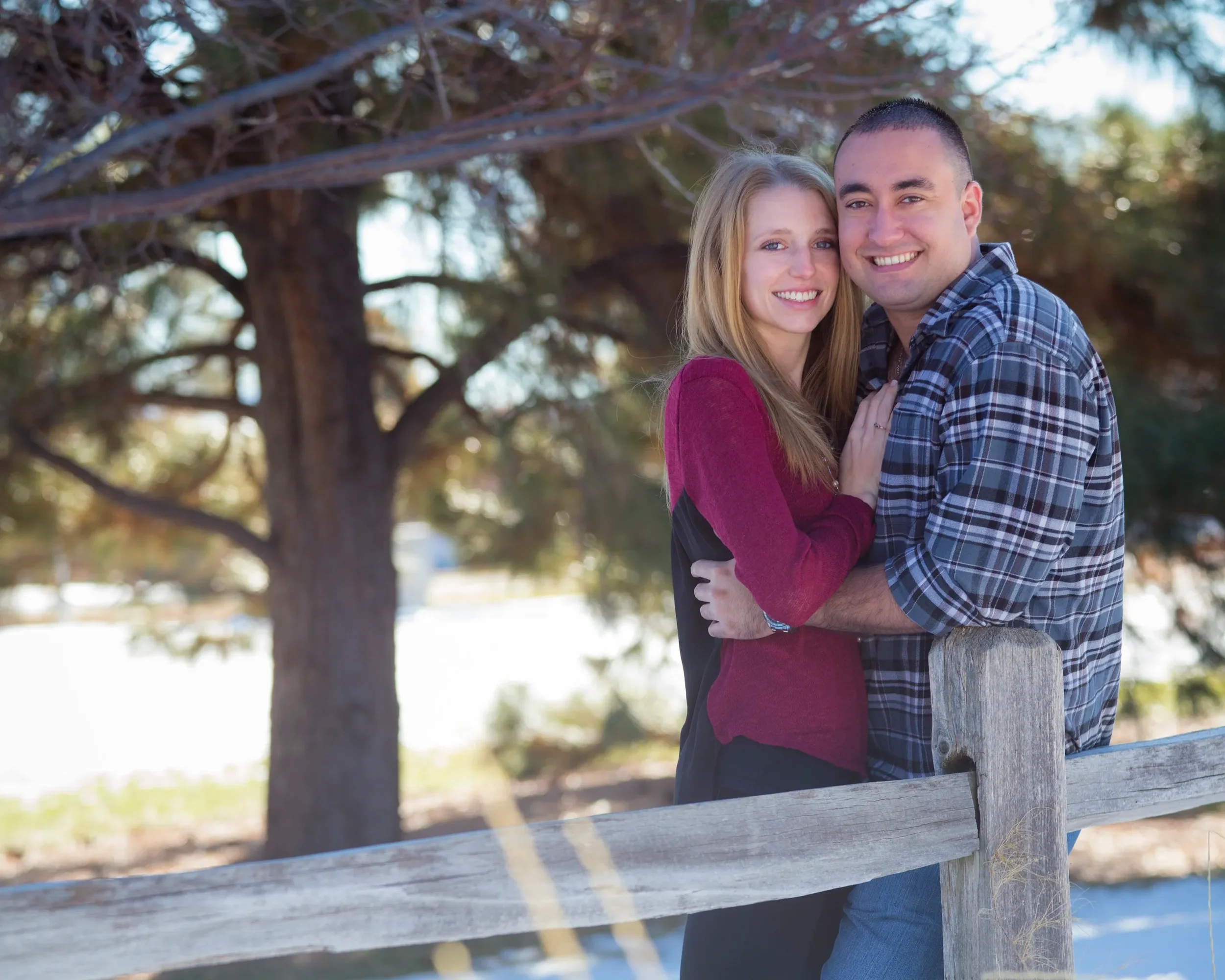 A smiling couple hugging outdoors near a wooden fence, with a large tree and greenery in the background.