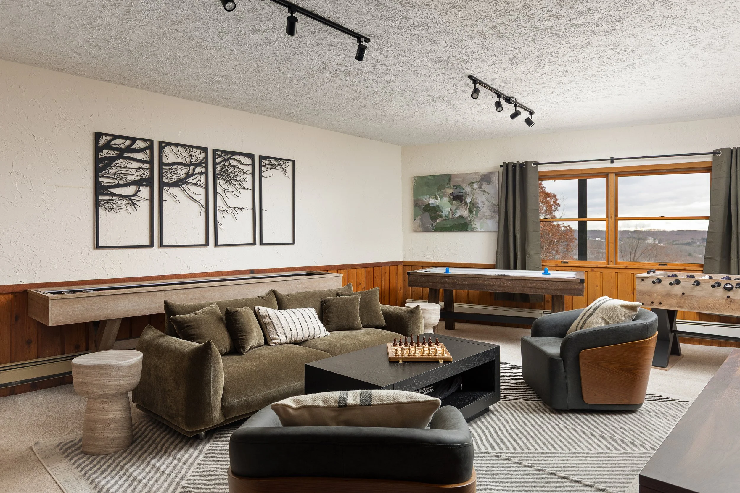 Living room with a green sofa, black coffee table with a chess set, a black and tan armchair, foosball table near the window, art on the white textured walls, and large window with green curtains overlooking a landscape.