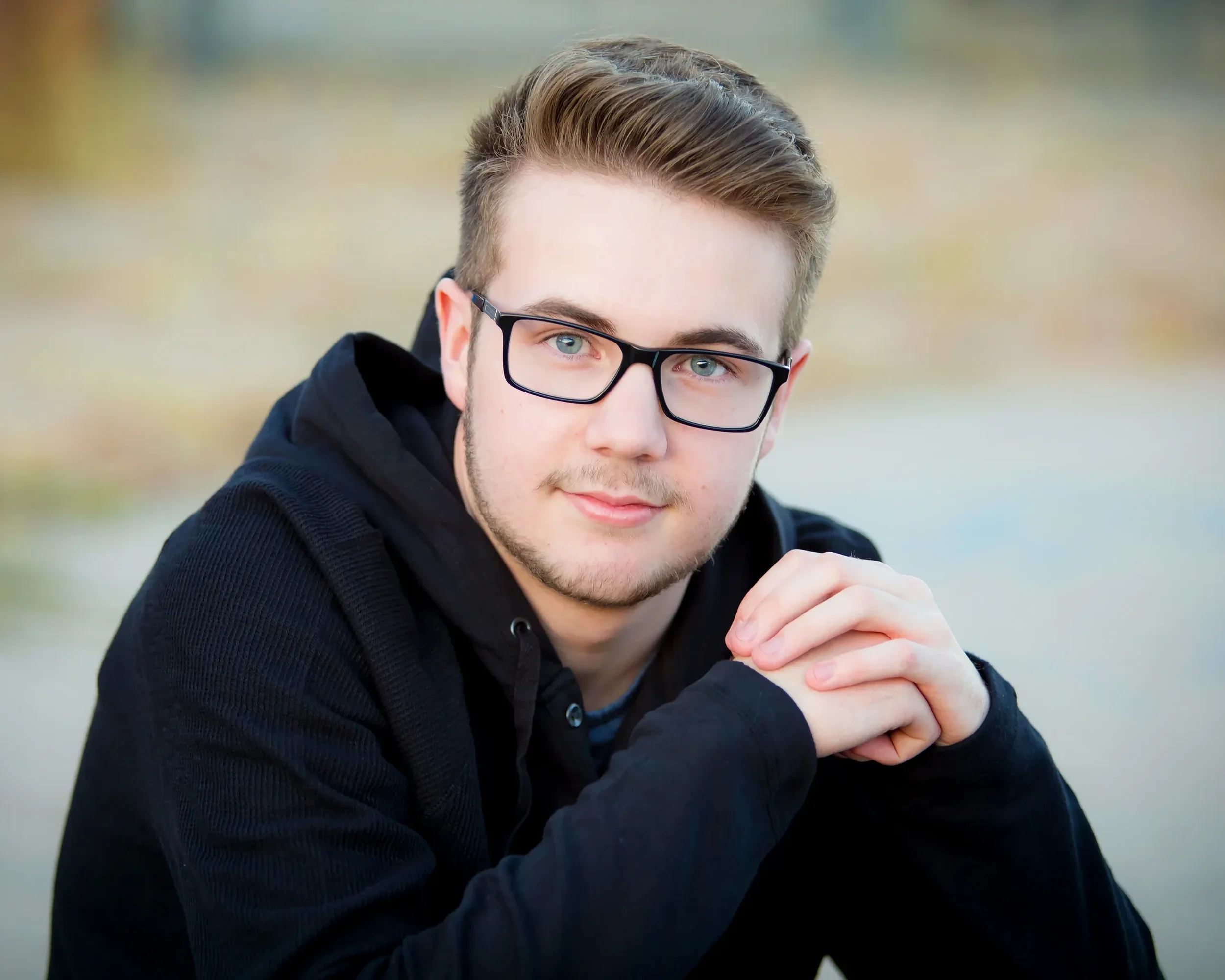 A young man with glasses, light brown hair, and a slight beard, wearing a black hoodie, is sitting outdoors with a blurred natural background, and is resting his chin on his hands.