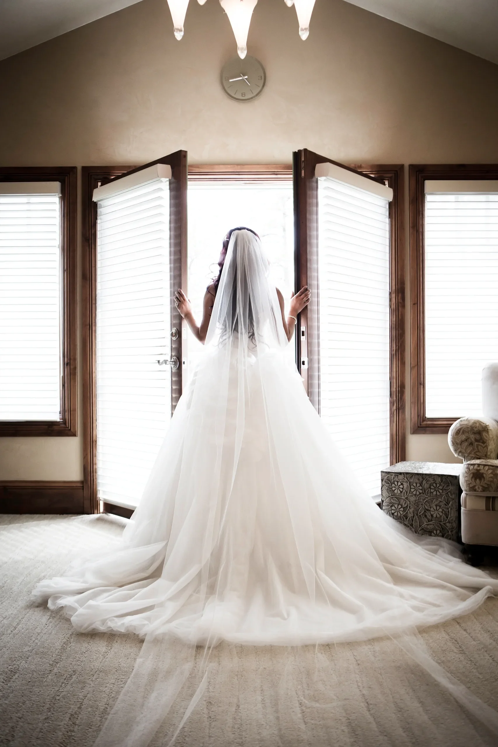 A bride in a white wedding dress and veil standing in front of open window shutters, looking outside, indoors with a clock on the wall above her.