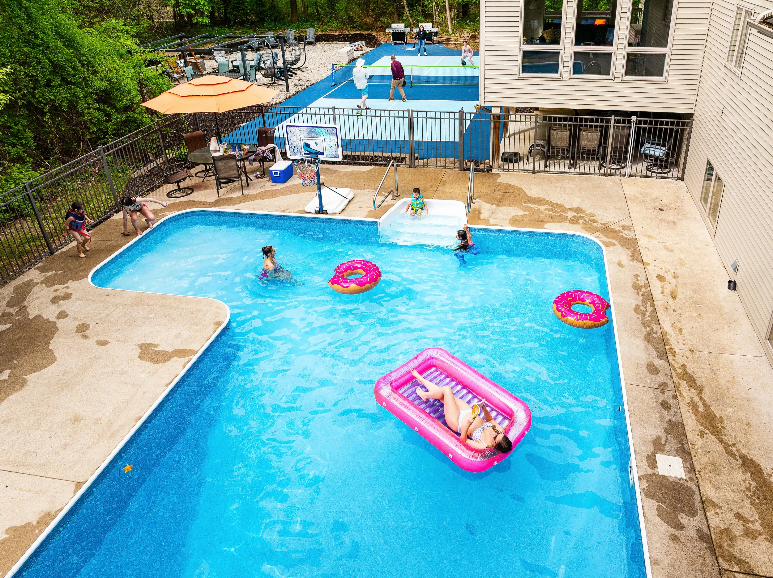 People enjoying a pool with some children playing and lounging on an inflatable float, with a swimming pool and a tennis court in the background surrounded by a fence and greenery.