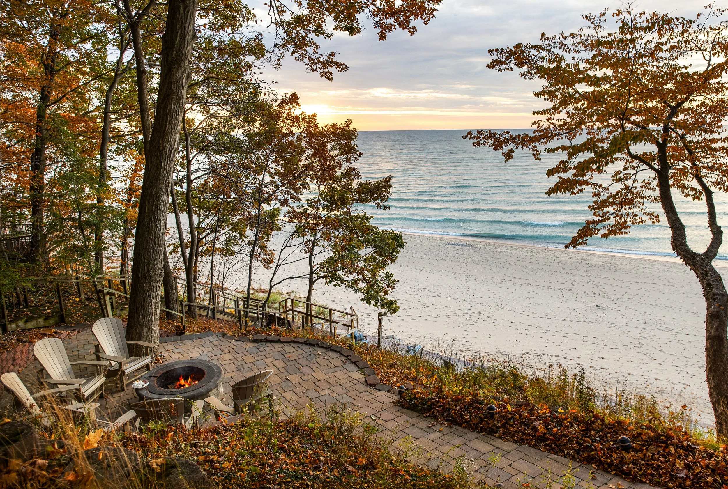 A beach scene during sunset viewed from a patio with Adirondack chairs and a fire pit surrounded by trees with autumn foliage.