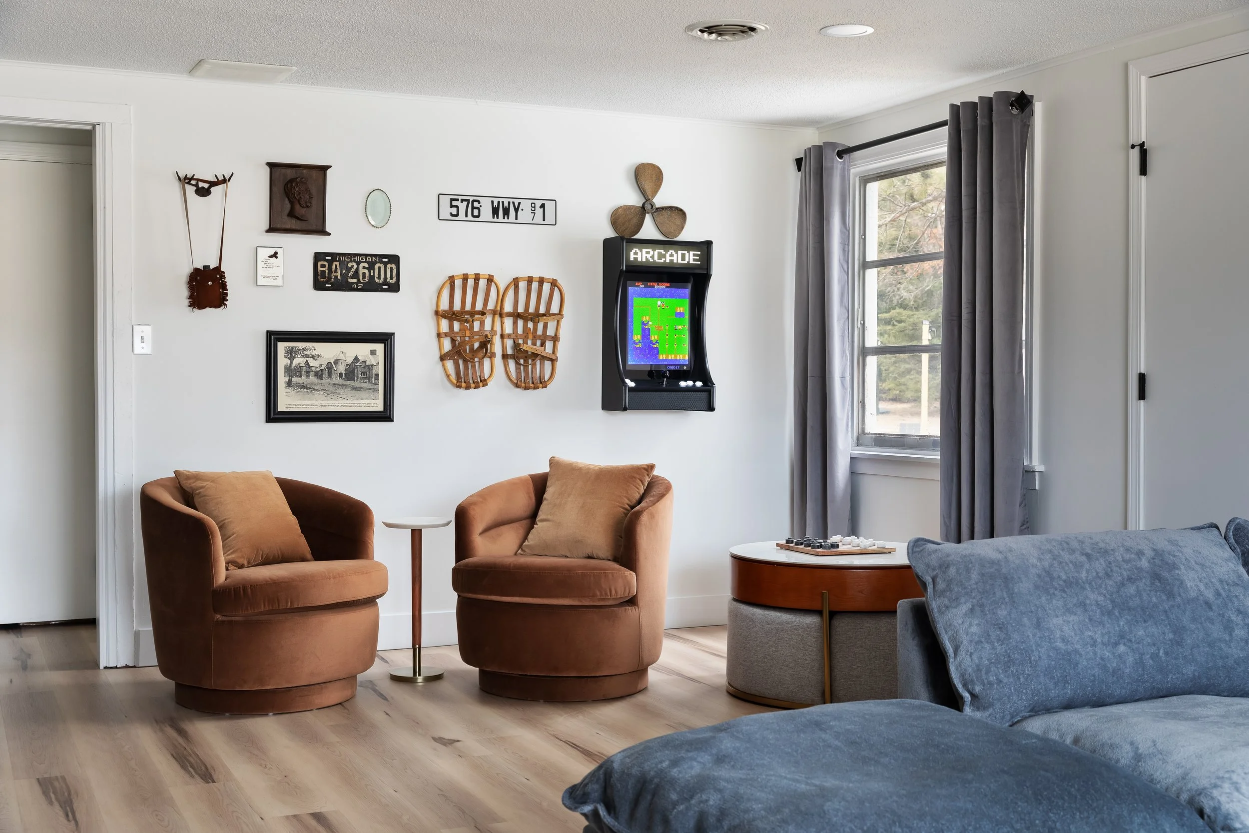 A cozy living room with two brown armchairs, a small white side table, and a gray sofa. The wall behind has various decorative items including vintage signs, a framed picture, and a small arcade game. A window with gray curtains lets in natural light