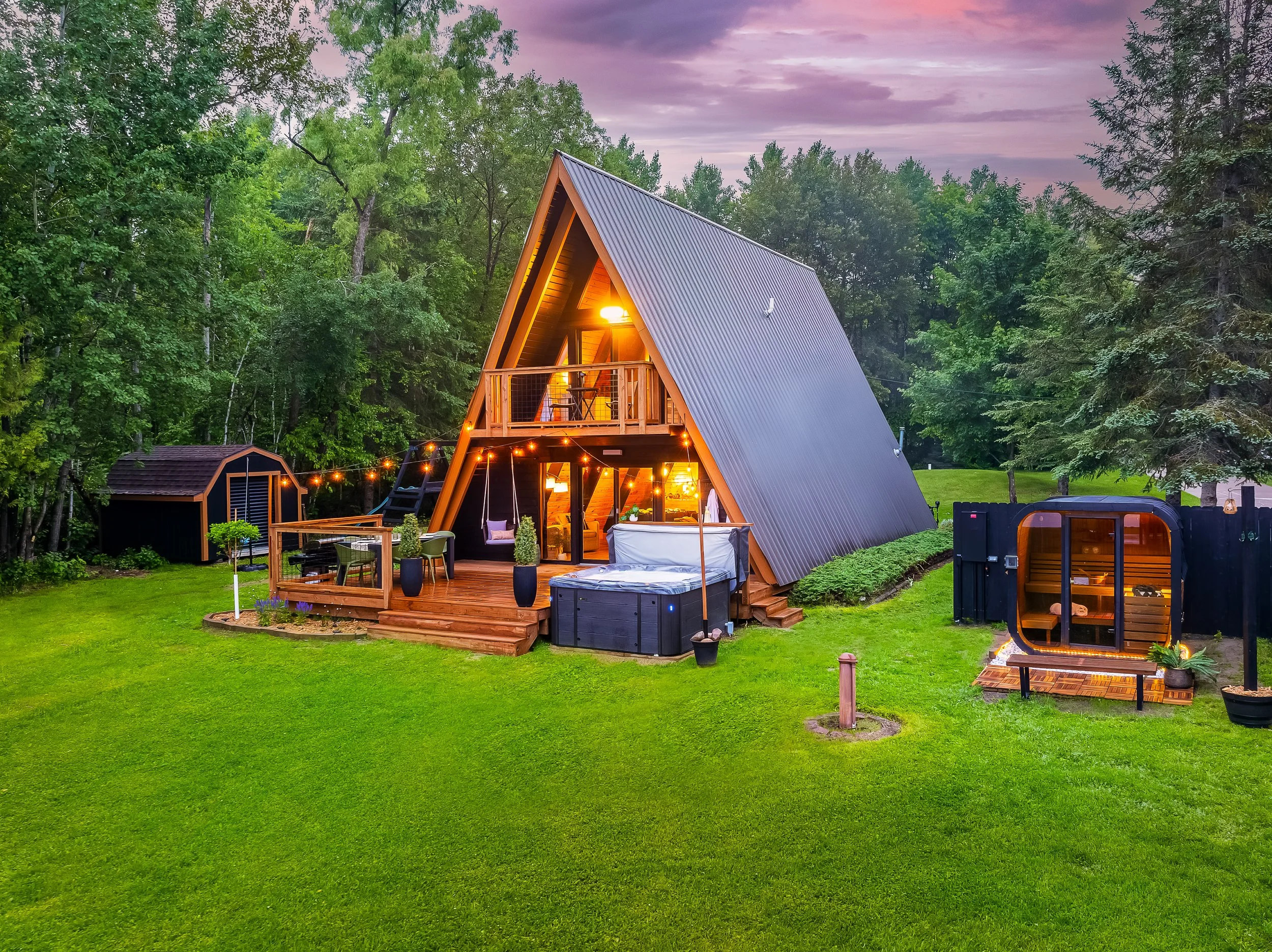 A modern A-frame cabin with a wooden deck, hot tub, and outdoor seating area, surrounded by green grass and trees
