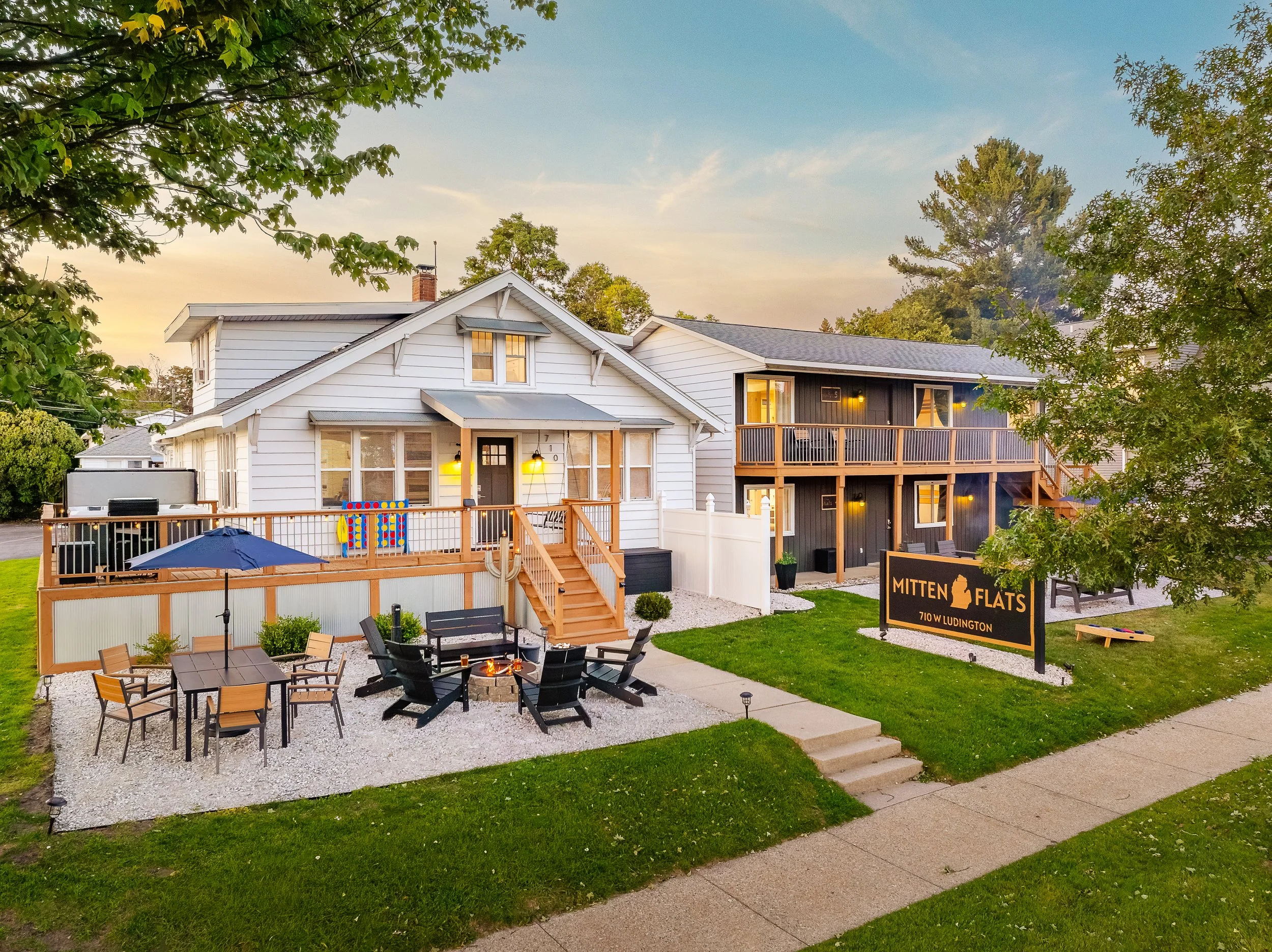 A two-story residential complex named 'Mitten Flats' with white and dark exterior. The property has a fenced back yard with outdoor seating, a table, and an umbrella. There are steps leading to the main entrance and a landscaped area with a sign and 