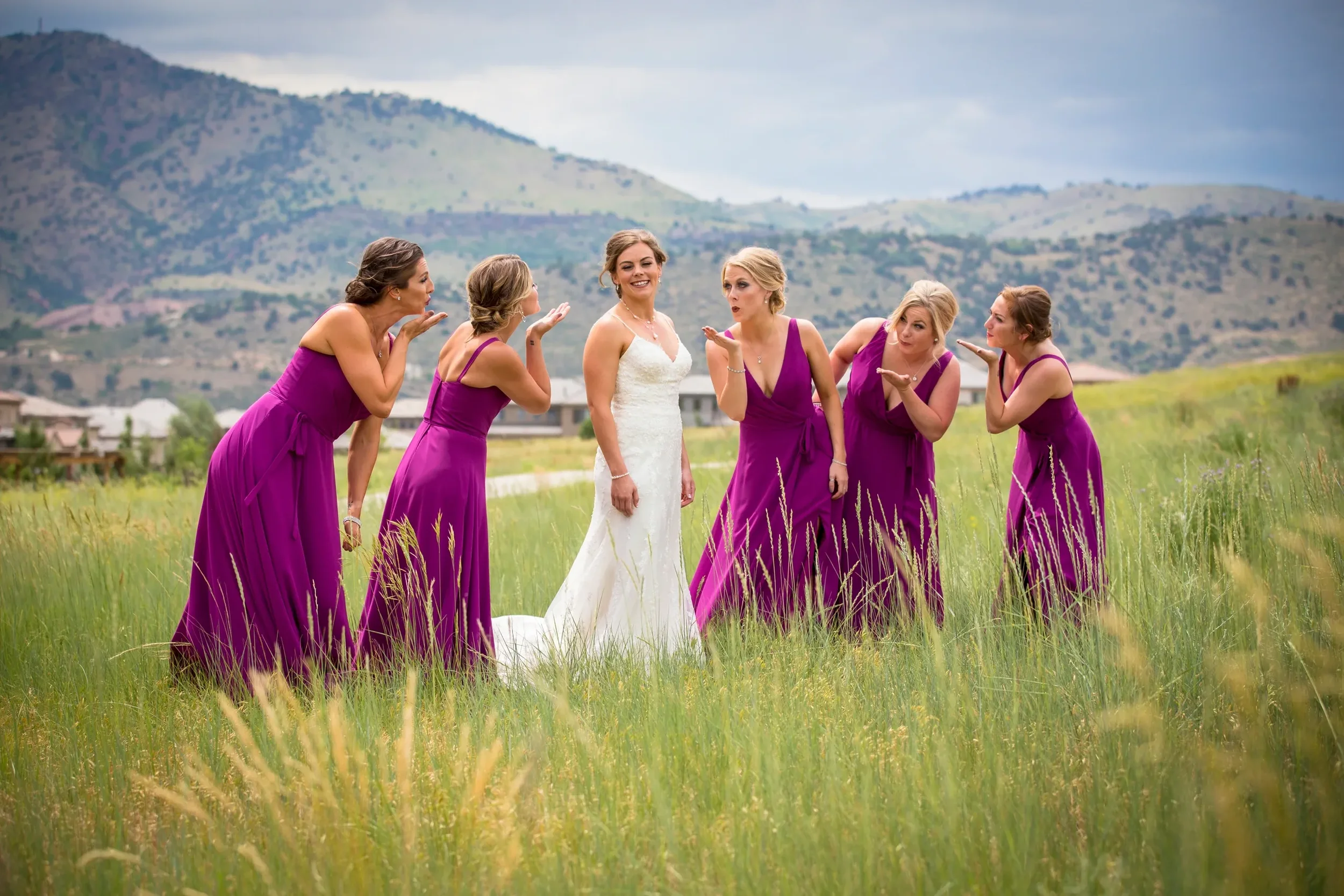 A bride in a white wedding dress stands in a field surrounded by five women in purple bridesmaid dresses. The women are blowing kisses towards the bride, who is smiling. The background features mountains and a cloudy sky.