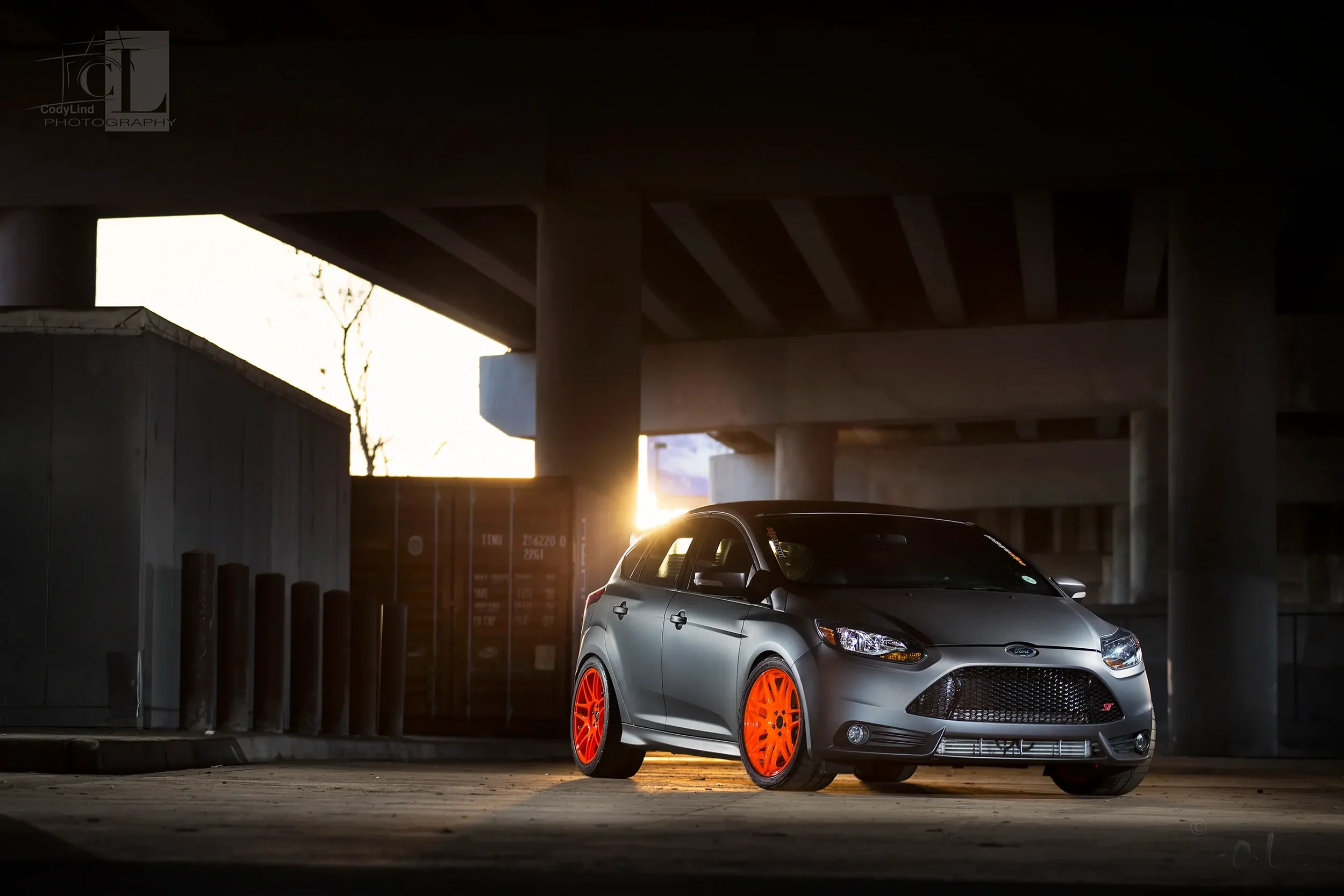 A gray hatchback car with bright orange rims parked under a highway overpass at sunset.