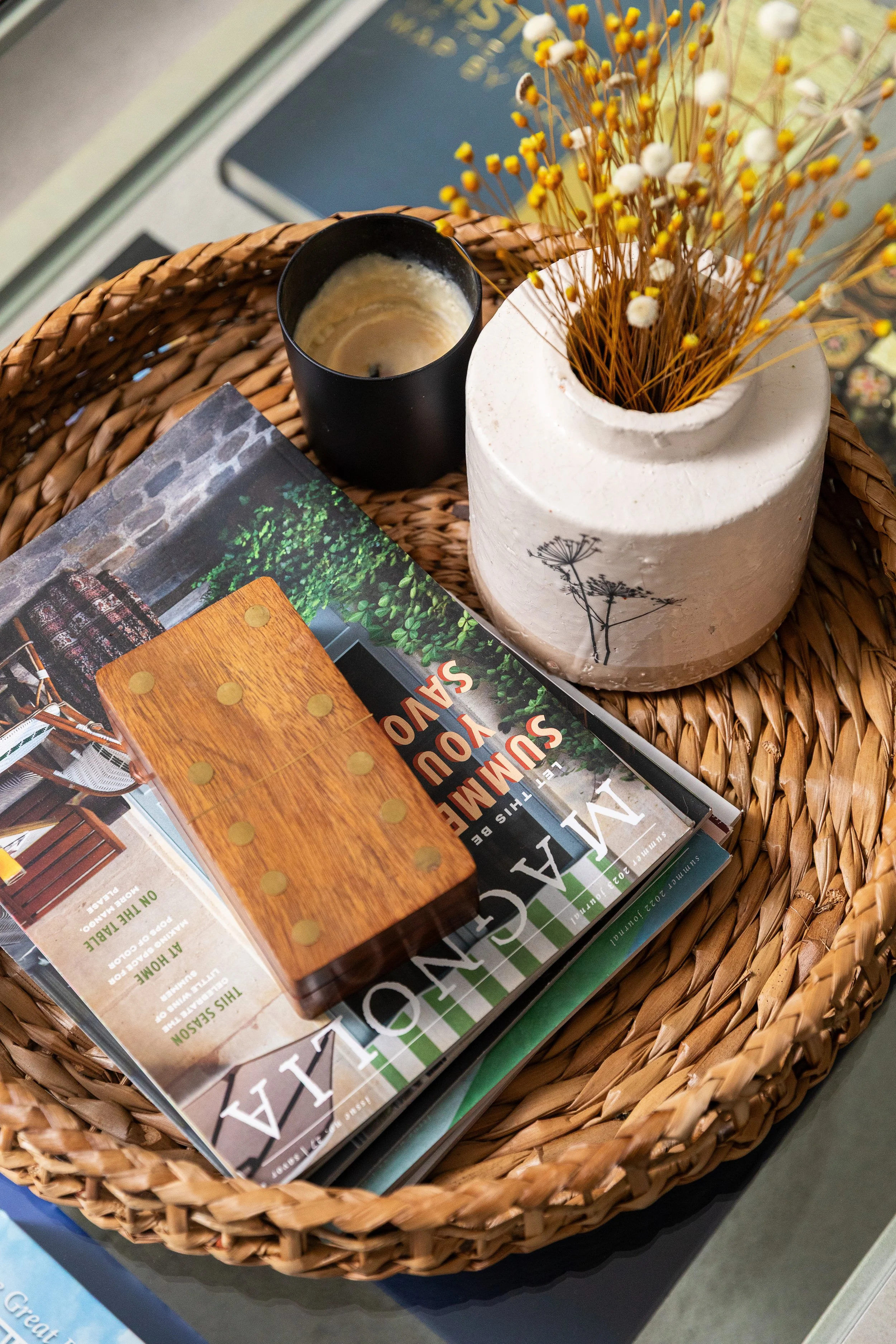 A wicker tray holding a magazine, a small black candle, and a cream-colored vase with dried flowers.