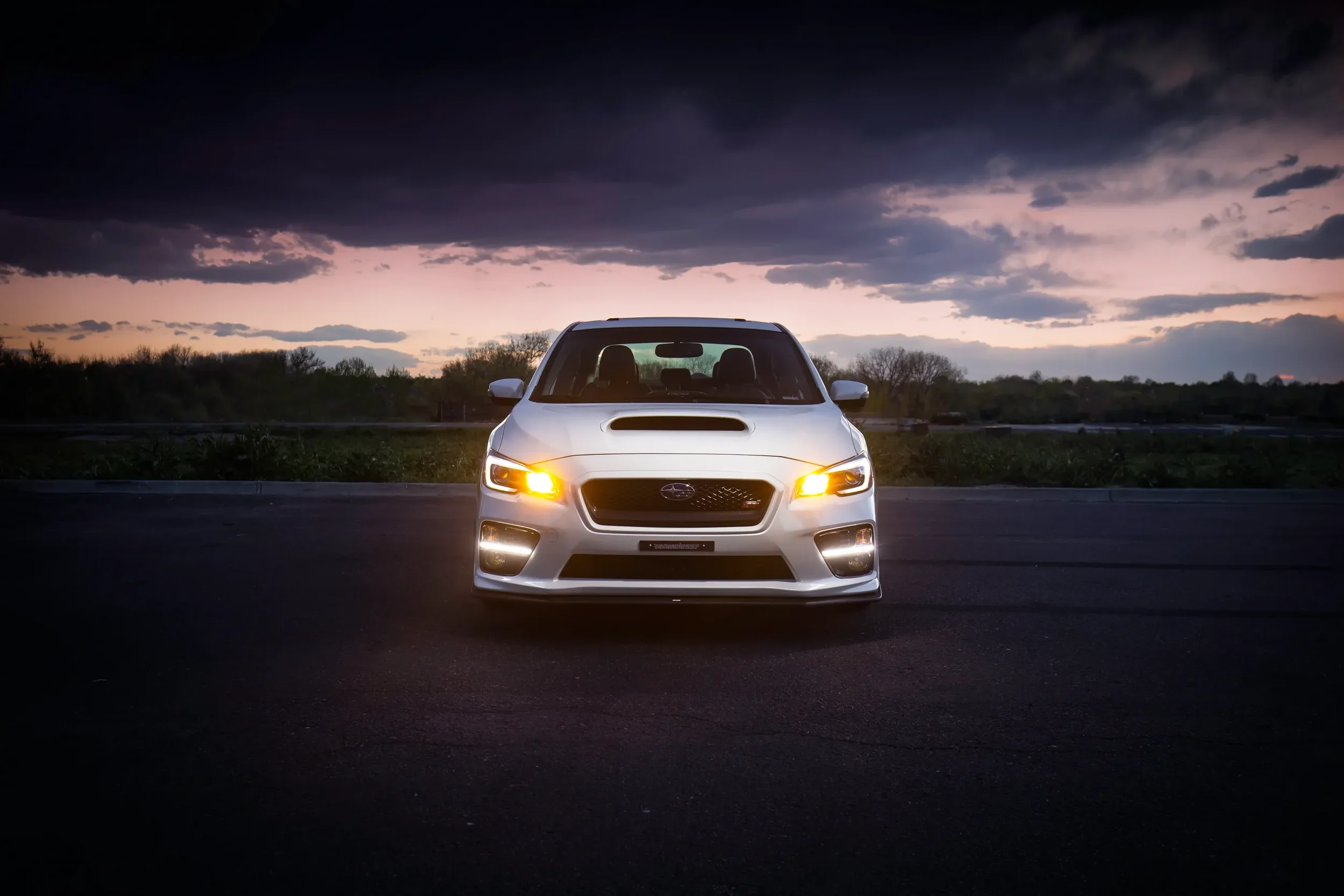 White Subaru car with headlights on parked on empty road at dusk with dark clouds and sunset in the background.