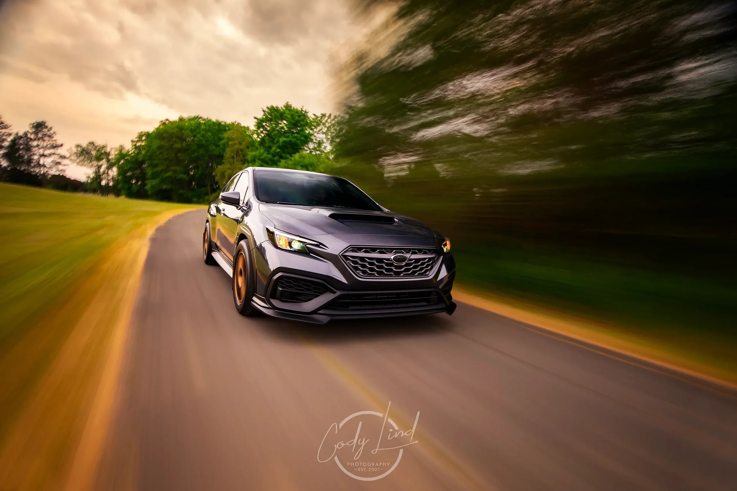 A black SUV with bronze wheels driving on a curved road through a park with trees, with a dramatic sky overhead.