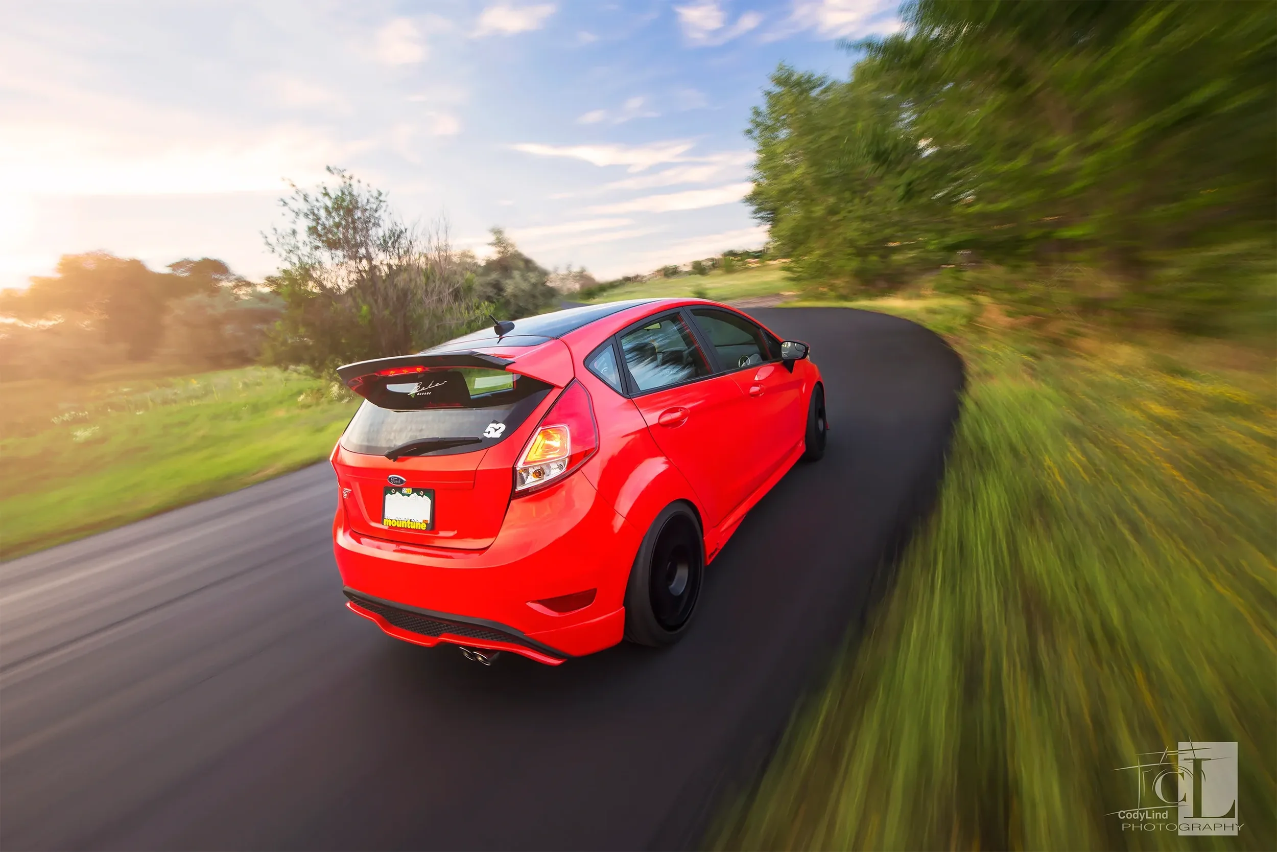 Red hatchback car driving on a winding road through green landscape with trees and grass, with motion blur indicating speed, during daytime with partly cloudy sky.