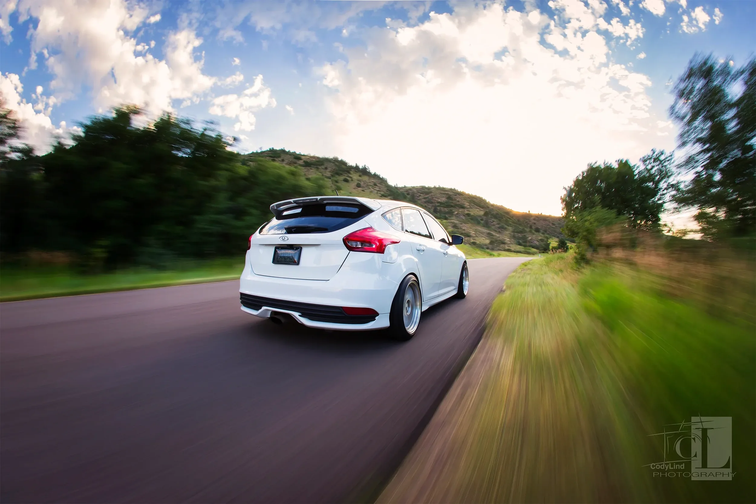 White hatchback car driving on a winding road through a landscape with green trees and hills during sunset.